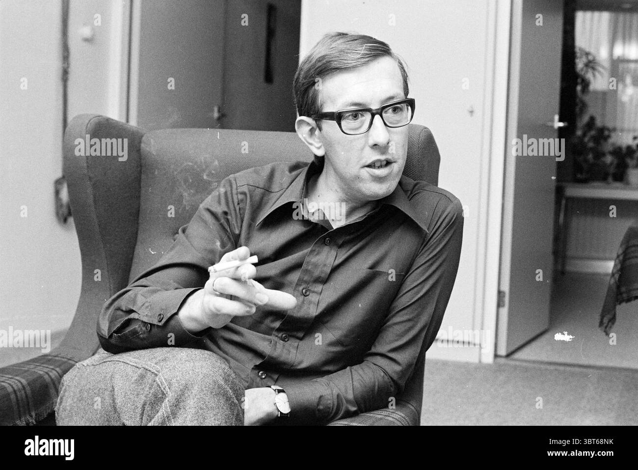Portrait Mr. City Portraits, Whizgle News, Dutch Desk, pays-Bas, 1950 - 2000 sur 08-08-1973. Ce sont les éléments de l'image. La scène capture un homme assis dans un fauteuil confortable et légèrement usé. Il a une expression contemplative, accentuée par ses lunettes et ses cheveux soigneusement peignés. Sa tenue se compose d'une chemise foncée et boutonnée et de pantalons gris, véhiculant une apparence chic et décontractée. Autour de lui, la pièce est légèrement éclairée, créant une atmosphère chaleureuse et accueillante. La lumière semble filtrer à partir d'une source invisible, projetant de douces ombres qui mettent en évidence les contours du furn Banque D'Images