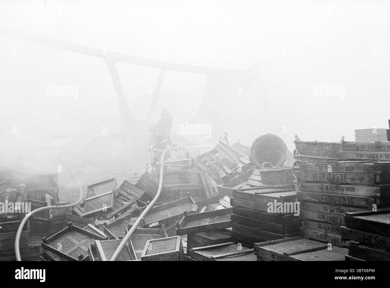 Hangar à ampoules à feu fa. Willemsen Hillegom Fire brigade Hillegom, Whizgle News, Dutch Desk, pays-Bas, 1950 - 2000 on 02-03-1980. Voici les rubriques de l'image. La scène capture un décor sombre et désolé, enveloppé dans une couche dense de brouillard qui imprègne l'air, créant une atmosphère éthérée. Au premier plan, un tas chaotique de caisses en bois est dispersé, leurs surfaces altérées faisant allusion à des années d'exposition aux éléments. Certaines caisses sont empilées précairement, tandis que d'autres sont renversées, suggérant une perturbation récente. Émergeant de la brume, des formes vagues peuvent être vues - Li Banque D'Images
