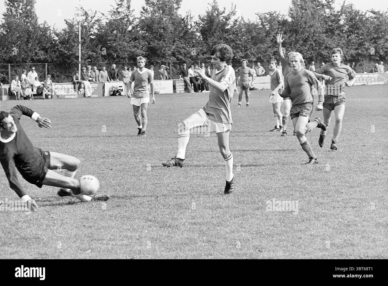 VSV - KFC Football V.S.V. Velsen Stormvogels, Whizgle News, Dutch Desk, pays-Bas, 1950 - 2000 le 04-09-1977. Ces rubriques apparaissent dans l'image. Dans une scène de football en plein air animée, deux équipes sont engagées dans un match animé sur un terrain vert dynamique. L'herbe semble fraîchement coupée, créant une toile de fond luxuriante pour l'action. Au premier plan, un joueur vêtu d'un maillot de couleur claire est prêt à tirer au but. Il est à mi-mouvement, une jambe levée et prête à se connecter avec le ballon. Son expression reflète une concentration intense. Le joueur est flanqué d'un gardien de but, vêtu d'une tenue sombre, qui Banque D'Images