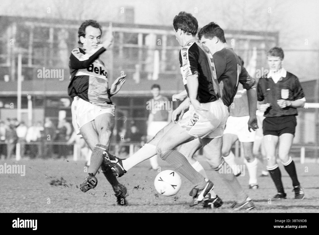 Stormvogels - DHC Football V.S.V. Velsen Stormvogels, Whizgle News, Dutch Desk, pays-Bas, 1950 - 2000 on 12-11-1989. Voici les rubriques de l'image. La scène se déroule sur un terrain herbeux, où un match de football dynamique est en cours. Les joueurs des deux équipes sont intensément concentrés, leurs expressions montrant la détermination et l'esprit de compétition. Un joueur, positionné sur la gauche, est capturé à mi-action, alors qu'il tente d'exécuter un coup de pied. Il porte un maillot avec un motif géométrique qui contraste avec les couleurs plus Unies de ses coéquipiers. Ses cheveux sont touffus, suggérant l'énergie du Banque D'Images