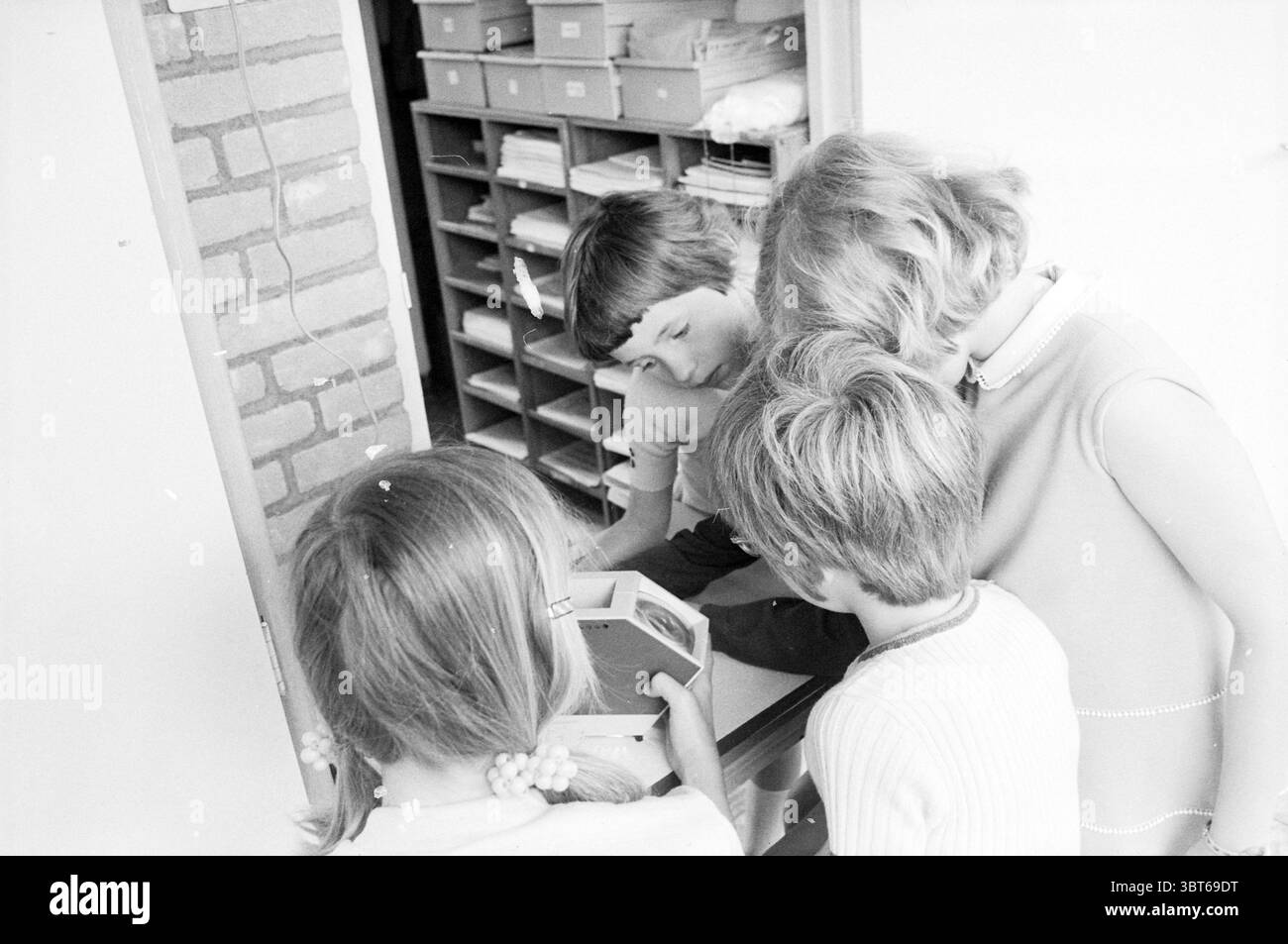 Report School Heemskerk Schools, Whizgle News, Dutch Desk, pays-Bas, 1950 - 2000 on 18-05-1971. Ces rubriques sont illustrées dans l'image. Dans un cadre intérieur lumineux, un petit groupe de quatre enfants se réunit autour d’une table, concentrés sur un objet carré placé devant eux. La salle est baignée de lumière naturelle, créant une atmosphère chaleureuse et accueillante. Les murs affichent une teinte douce, tandis que le sol a un design simple qui complète l'ambiance globale. Les enfants font preuve d'un mélange de curiosité et d'excitation. Deux garçons et deux filles sont blottis ensemble, leurs expressions véhiculant Banque D'Images