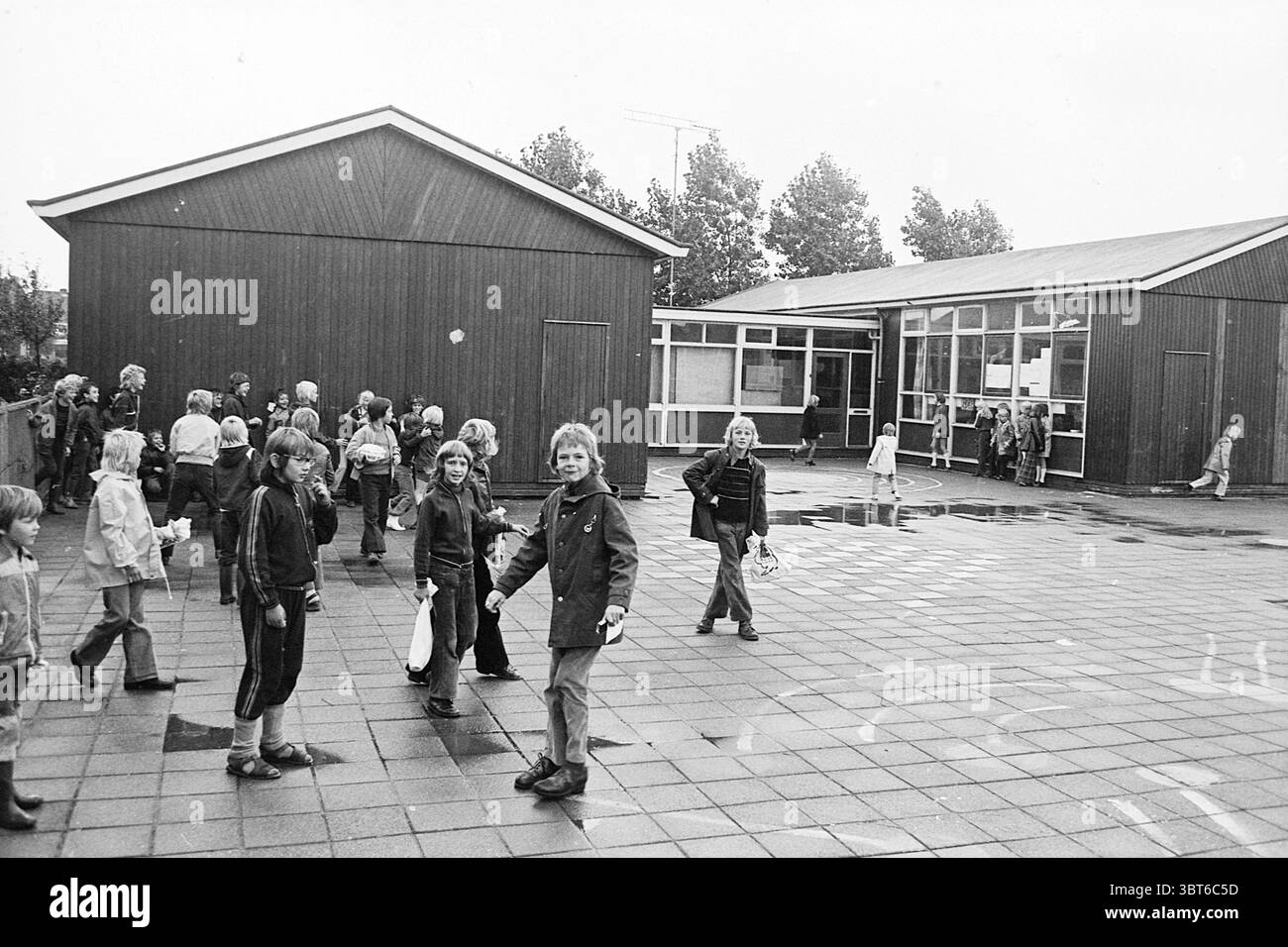 Terrain de jeu Mearten van Heemskerckschool. Terrains de jeux et écoles de terrains de jeux, Whizgle News, Dutch Desk, pays-Bas, 1950 - 2000 on 09-09-1974. L'image montre ces rubriques. Dans une scène représentant une cour d'école, un groupe diversifié d'enfants et d'adultes est réuni, créant une atmosphère animée. Le décor présente deux structures en bois au design simple et utilitaire, caractérisées par un revêtement brun foncé. Ces bâtiments sont fonctionnels, avec de nombreuses fenêtres qui reflètent un ciel légèrement couvert. Le sol est fait de grandes tuiles carrées, certaines scintillantes de la pluie récente, améliorant le s. Banque D'Images