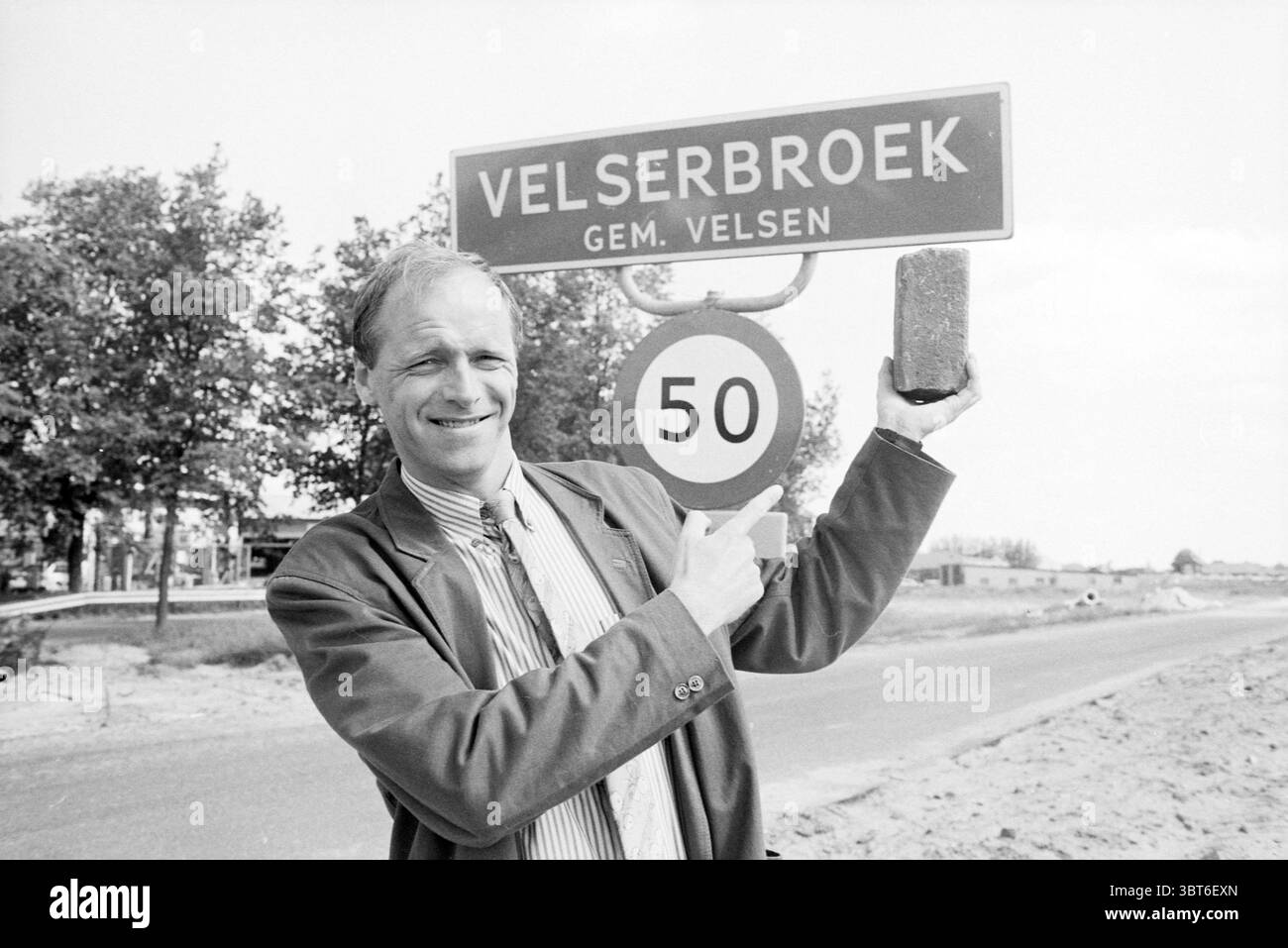 Mr. Hoogeveen au panneau Velserbroek Personen Velserbroek, Whizgle News, Dutch Desk, pays-Bas, 1950 - 2000 le 27-05-1989. L'image montre ces rubriques. Un homme se tient sur le bord d'une route, mis en évidence par un ciel ensoleillé qui projette une ambiance lumineuse et joyeuse sur la scène. Il est habillé d'un costume, qui semble légèrement formel contre le cadre extérieur décontracté. L'homme a un comportement amical, avec un large sourire alors qu'il pointe avec enthousiasme vers un panneau de signalisation. Le panneau de signalisation présente une forme circulaire proéminente avec un chiffre en gras « 50 » en son centre, indiquant une limite de vitesse, sous Whic Banque D'Images