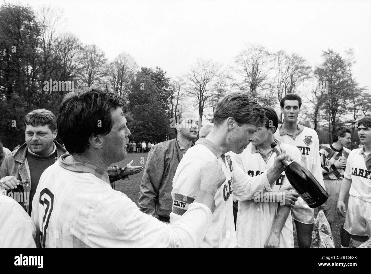 Bloemendaal champion, Whizgle News, Dutch Desk, pays-Bas, 1950 - 2000 sur 04-05-1991. Ces rubriques apparaissent dans l'image. La scène montre un groupe d'individus jubilants célébrant après un événement sportif, probablement un match de football, dans un cadre extérieur. Les joueurs sont vêtus de maillots blancs et foncés rayés, ces caractéristiques mettent en valeur les couleurs de l'équipe. Certains d’entre eux ont des expressions festives, caractérisées par des sourires et des gestes animés, reflétant un sentiment collectif de réussite et de bonheur. Au premier plan, un individu se distingue, tenant une grande bouteille de champagne, son sur brillant Banque D'Images