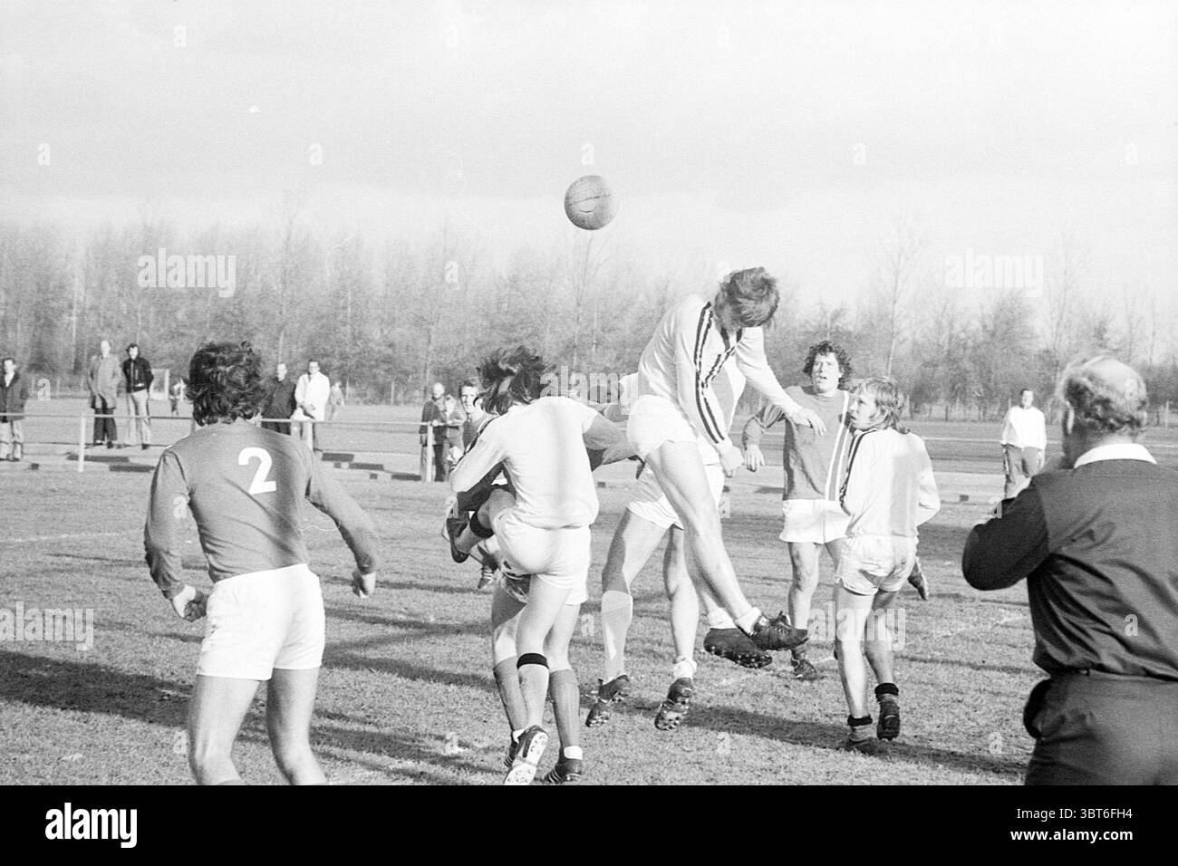 Match de football, Whizgle News, Dutch Desk, pays-Bas, 1950 - 2000 en 1973. Ces rubriques apparaissent dans l'image. La scène présente un moment dynamique sur un terrain herbeux, où un groupe d'athlètes est engagé dans un jeu de rugby animé. Au premier plan, un joueur saute en l'air, les bras tendus, tentant d'attraper un ballon. Son uniforme est majoritairement blanc, avec quelques rayures contrastées. Les couleurs contrastées des uniformes portés par les autres joueurs, certains en rouge et d'autres en blanc, créent un contraste visuel éclatant avec l'herbe verte luxuriante en dessous. Autour de ces joueurs, le Banque D'Images