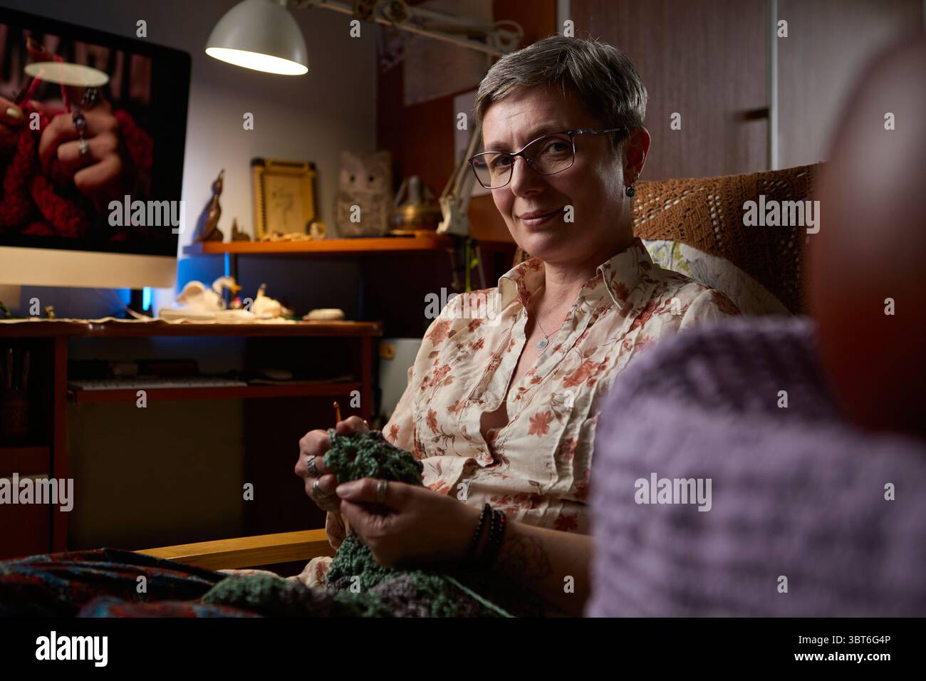 Une femme mature confiante assise dans un environnement domestique confortable, crochetant une pièce faite à la main. L'éclairage chaleureux et le cadre détendu Banque D'Images