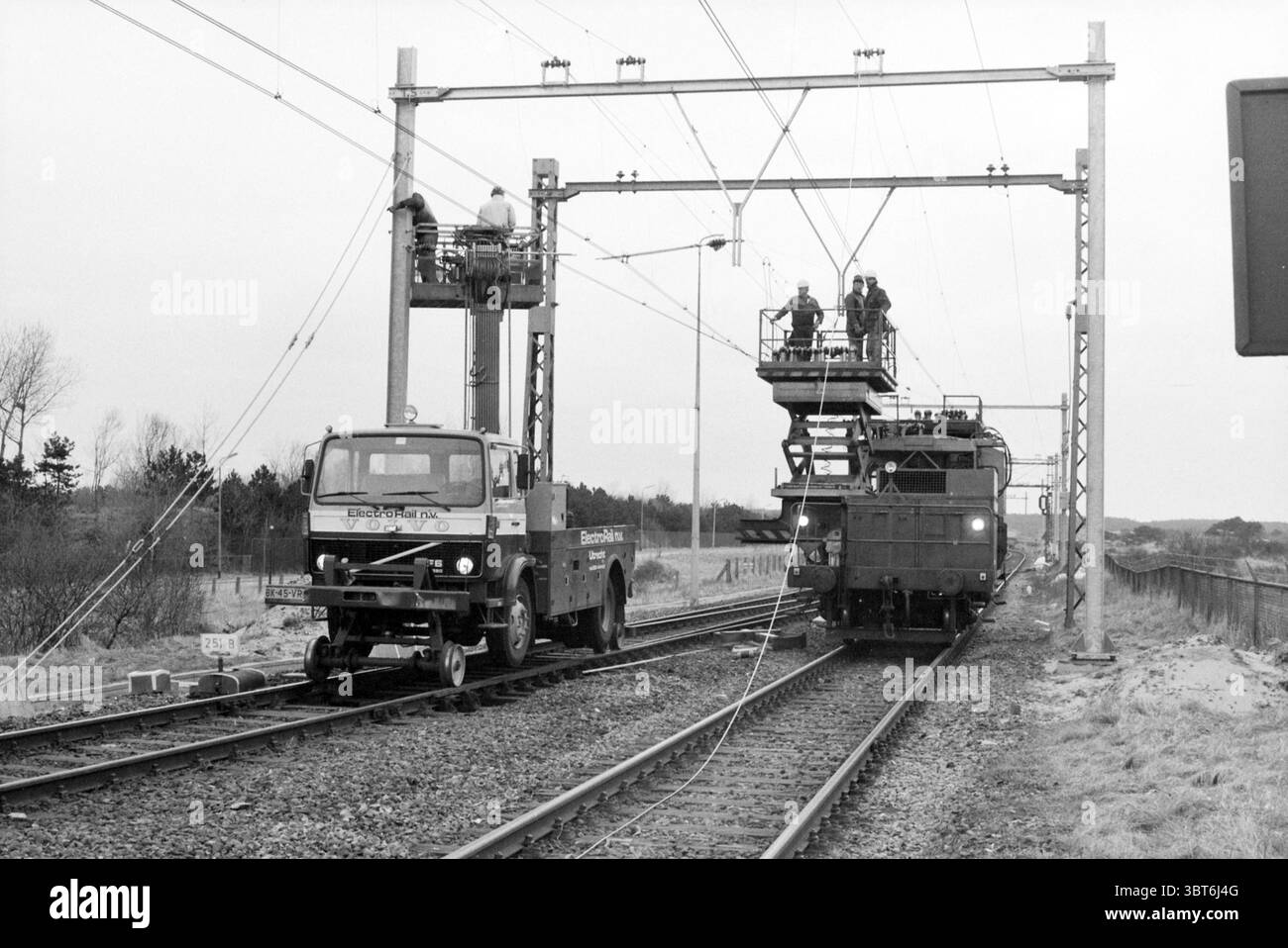 Travaux sur la ligne ferroviaire de Zandvoort chemins de fer Zandvoort, Whizgle News, Dutch Desk, pays-Bas, 1950 - 2000 le 04-04-1986. L'image contient ces rubriques. La scène représente un décor ferroviaire, caractérisé par une palette monochromatique qui souligne les nuances de gris. Deux véhicules sont bien positionnés sur les voies. À gauche, un camion utilitaire de construction robuste est stationné, mettant en valeur sa conception fonctionnelle. Son corps est gris foncé ou éventuellement noir, contrastant avec les tons plus clairs qui l'entourent. À droite, un train électrique dégage un sentiment de puissance industrielle, avec des lignes audacieuses et Banque D'Images