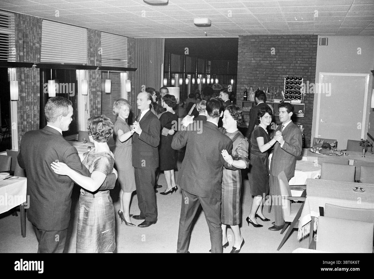 Soirée dansante et amusante., Whizgle News, Dutch Desk, pays-Bas, 1950 - 2000. L'image montre ces rubriques. La scène représente un rassemblement animé dans un cadre de restaurant moderne du milieu du siècle. L'intérieur présente un mélange de tons chauds et froids, mis en évidence par des panneaux en bois sur les murs et des surfaces polies. Une série de luminaires doux et d'ambiance suspendus au plafond, projetant une douce lueur qui améliore l'atmosphère intime. Au centre, un groupe de couples élégamment habillés dansent de près, incarnant un sentiment de joie et de célébration. Les femmes portent des robes élégantes et ajustées qui Banque D'Images