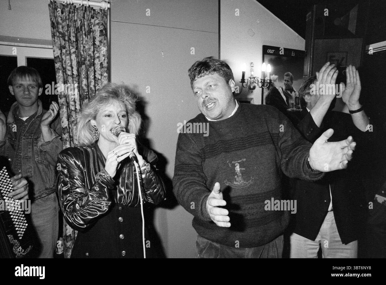 Bachelor Night Alex Freijser, Whizgle News, Dutch Desk, pays-Bas, 1950 - 2000 le 17-03-1991. Ces rubriques apparaissent dans l'image. La scène capture un rassemblement animé dans un cadre intérieur. Au centre, une femme aux cheveux blonds volumineux et stylés chante passionnément dans un microphone, exsudant enthousiasme et confiance. Elle porte une veste noire brillante avec des épaulettes distinctes, soulignant un choix de mode audacieux typique d'une époque dynamique. À sa droite, un homme chante animée, fait des gestes enthousiastes des deux mains, créant une atmosphère engageante. Il est sombre, touffu Banque D'Images