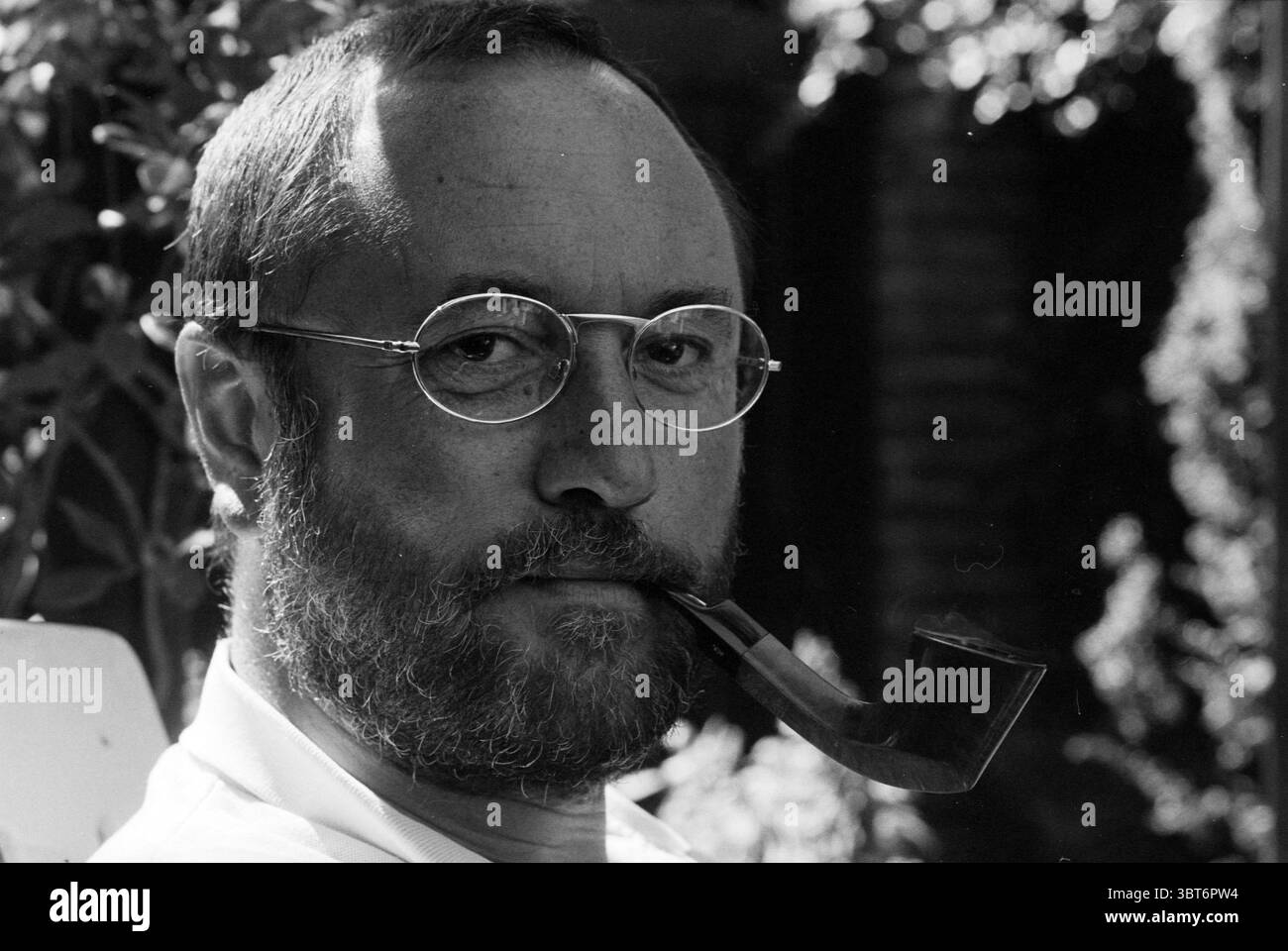 Henk van Dorp Pipe Smoking World Championships, Whizgle News, Dutch Desk, pays-Bas, 1950 - 2000 le 16-08-1993. Ces rubriques apparaissent dans l'image. La scène met en scène un homme avec une expression réfléchie, assis dans un cadre extérieur serein. Il a une barbe pleine et porte des lunettes rondes qui captent la lumière subtilement. Une longue pipe à fumer repose confortablement entre ses lèvres, lui donnant un comportement contemplatif. L'arrière-plan est rempli de verdure luxuriante, suggérant un jardin ou un parc, avec des ombres douces et tapissées jouant sur son visage. La lumière du soleil filtre à travers les feuilles, créant une chaleur et une tonicité Banque D'Images