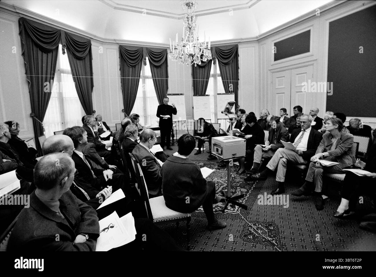 Réunion de travail du conseil municipal + conseil, Whizgle News, Dutch Desk, pays-Bas, 1950 - 2000 le 14-02-2000. L'image montre ces rubriques. La scène se déroule dans une salle élégamment décorée, caractérisée par ses hauts plafonds et son éclairage doux et chaleureux qui crée une atmosphère accueillante. Des rideaux riches et sombres encadrent les grandes fenêtres, permettant à la lumière naturelle de filtrer, contribuant ainsi à l'ambiance. Un grand lustre orné pend au plafond, projetant une douce lueur à travers l'espace. Au premier plan, un groupe d'individus est assis autour de la pièce, disposés en cercle informel. Le Banque D'Images