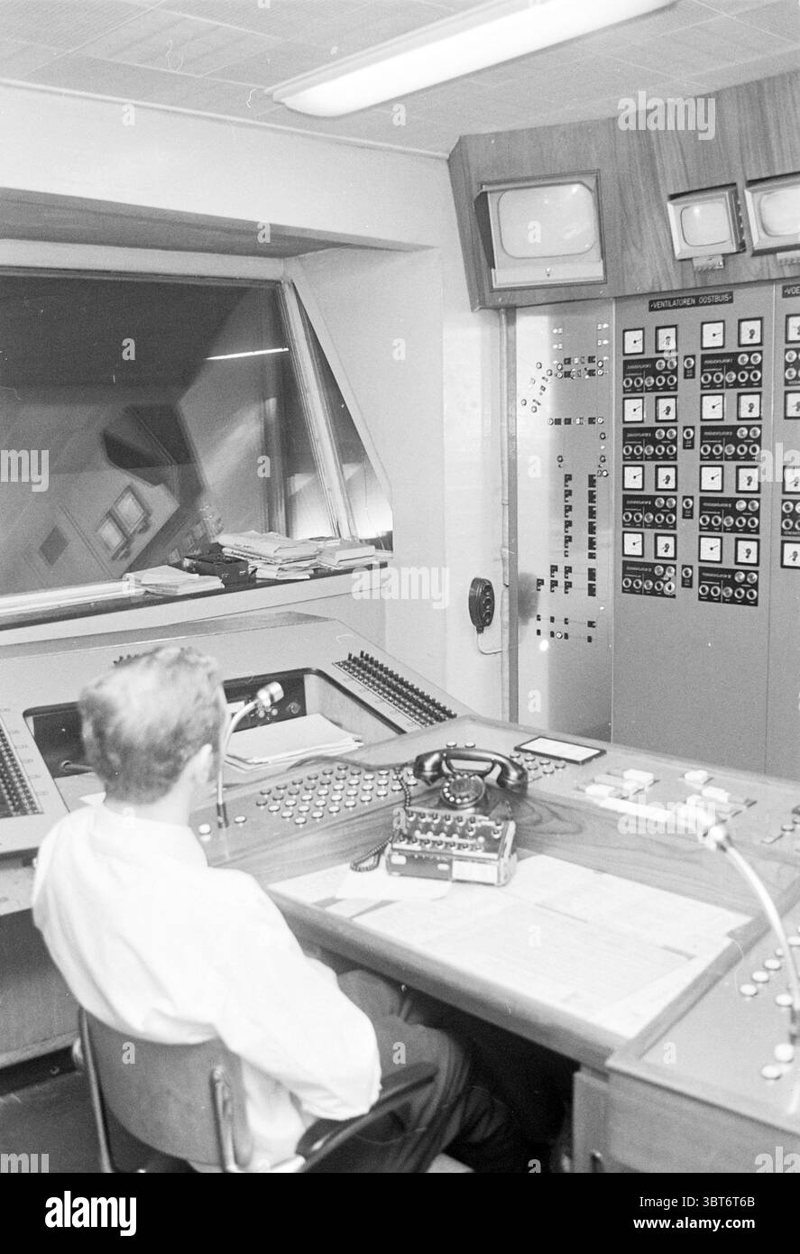 Salle de contrôle., Whizgle News, Dutch Desk, pays-Bas, 1950 - 2000. L'image montre ces rubriques. La scène représente une salle de contrôle vintage, caractérisée par son design du milieu du XXe siècle. Un individu solitaire est assis à un grand bureau incurvé, qui est équipé d'un ensemble de boutons, de cadrans et de leviers, suggérant un système complexe en fonctionnement. L'individu, vêtu d'une chemise formelle, fait face à une grande fenêtre en verre qui offre une vue de l'espace au-delà, bien que les détails de cet espace ne soient pas visibles. Le pupitre de commande a une surface en bois poli avec divers instruments stratégiques Banque D'Images