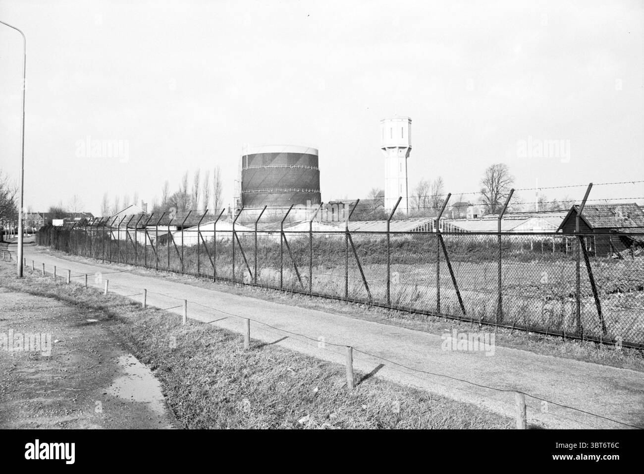 Nurserie sur le Cruquiusweg Haarlem aperçu. Cultivateurs de pépinières Haarlem Cruquiusweg pays-Bas, Whizgle News, Dutch Desk, pays-Bas, 1950 - 2000 on 12-11-1963. L'image inclut ces rubriques. La scène dépeint un paysage industriel sombre caractérisé par un sentiment de désolation. Au premier plan, un chemin étroit longe une clôture périmétrique surmontée de barbelés, laissant présager un accès restreint. Le chemin est bordé d'herbe qui semble dépouillée et unkemptée, traquant la voie à suivre. Sur le côté, une grande structure cylindrique, probablement un réservoir de stockage, se tient en évidence. Il est peint i. Banque D'Images