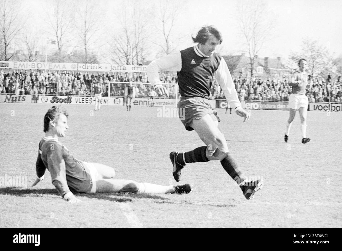 Haarlem - Excelsior Football Haarlem, Whizgle News, Dutch Desk, pays-Bas, 1950 - 2000 le 04-05-1975. Ces rubriques sont illustrées dans l'image. La scène capture un moment sur un terrain de football animé, rempli d'énergie et de mouvement. L'herbe est vibrante et bien entretenue, contrastant avec le ciel bleu vif ci-dessus, qui indique un temps clair. Au premier plan, un joueur est attrapé à mi-foulée, portant un uniforme sombre avec un motif rayé, mettant en valeur un mélange de couleurs qui transmettent un sentiment de vigueur et de concentration. Son expression est intense alors qu'il manœuvre devant un autre joueur, qui est sur le gr Banque D'Images