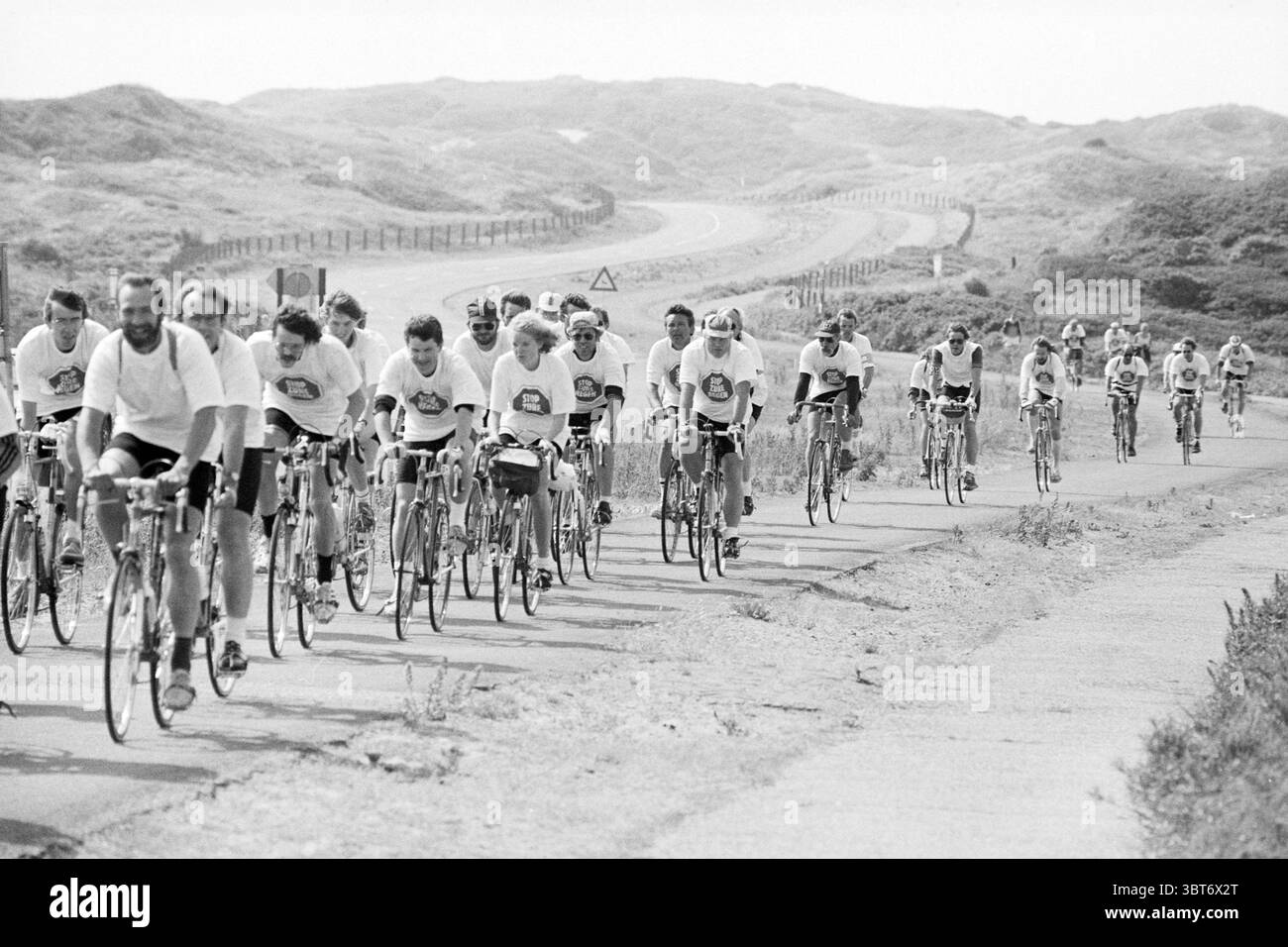 Vrom pistes cyclables cyclables cyclistes, Whizgle News, Dutch Desk, pays-Bas, 1950 - 2000 le 21-06-1986. Ce sont les éléments de l'image. Un groupe de cyclistes est vu naviguer sur une route sinueuse qui s'étend à travers un paysage doucement vallonné. La scène est majoritairement monochrome, ce qui lui confère une qualité nostalgique et intemporelle. Au premier plan, un groupe de cyclistes mène le peloton, tous portant des chemises blanches assorties ornées d’un emblème circulaire, suggérant un sentiment d’unité ou d’esprit d’équipe. Leurs vélos varient légèrement dans la conception, mais tous semblent bien utilisés, reflétant le de de leurs propriétaires Banque D'Images
