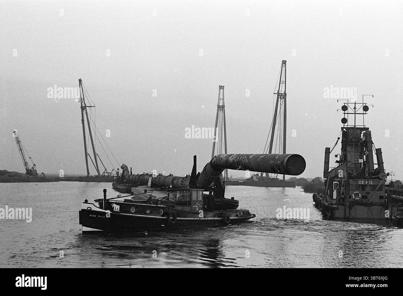 Pose de platine Drachten J.G. Nelis IJmuiden, Whizgle News, Dutch Desk, pays-Bas, 1950 - 2000 le 07-06-1971. L'image inclut ces rubriques. La scène représente une journée grise et nuageuse près d'une rivière ou d'un canal, donnant un sentiment de calme mais d'humeur légèrement sombre. Au premier plan, un modeste remorqueur navigue dans l'eau, sa coque sombre contrastant avec les tons sourds de l'environnement. L'eau apparaît immobile, reflétant la douce lumière du ciel. Derrière le remorqueur, un gros tuyau s'étend du navire, allant vers un chantier de construction industrielle. La surface du tuyau est sombre et usée, indic Banque D'Images