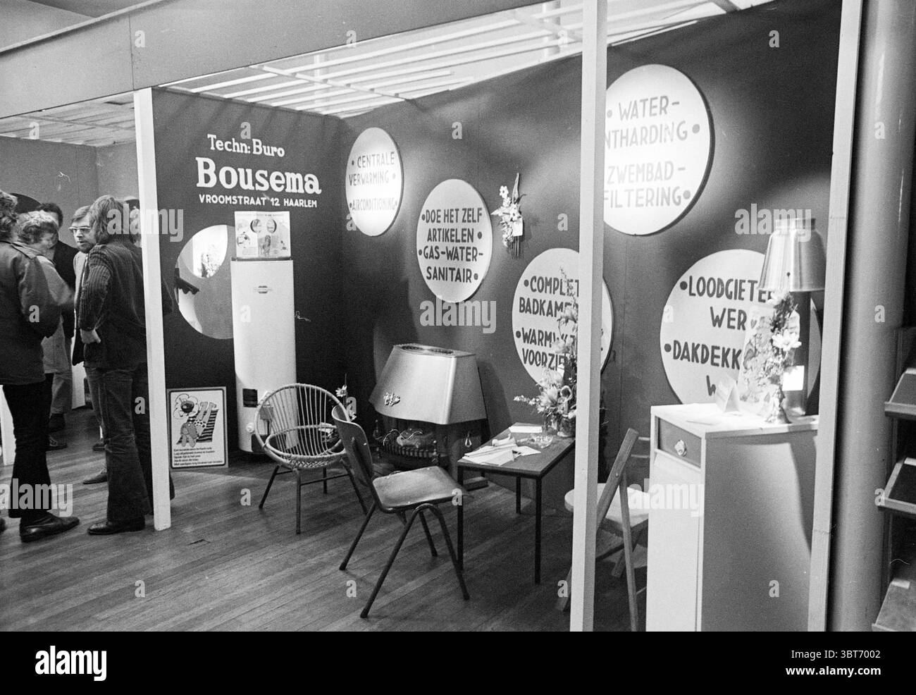 Cuisinière cuisines et sanitaires Fair., Whizgle News, Dutch Desk, pays-Bas, 1950 - 2000. Ce sont les éléments de l'image. La scène représente un espace d'exposition intérieur caractérisé par une esthétique élégante et moderne. Les murs sont peints d'un noir profond, avec des affichages ronds et de couleur claire qui mettent en valeur diverses offres de produits. Ces éléments contribuent à un fort contraste, rendant les écrans visuellement frappants. À gauche, un petit groupe de personnes est engagé dans la conversation, leur tenue suggérant une atmosphère décontractée, mais légèrement formelle. Ils semblent discuter de diverses aspec Banque D'Images