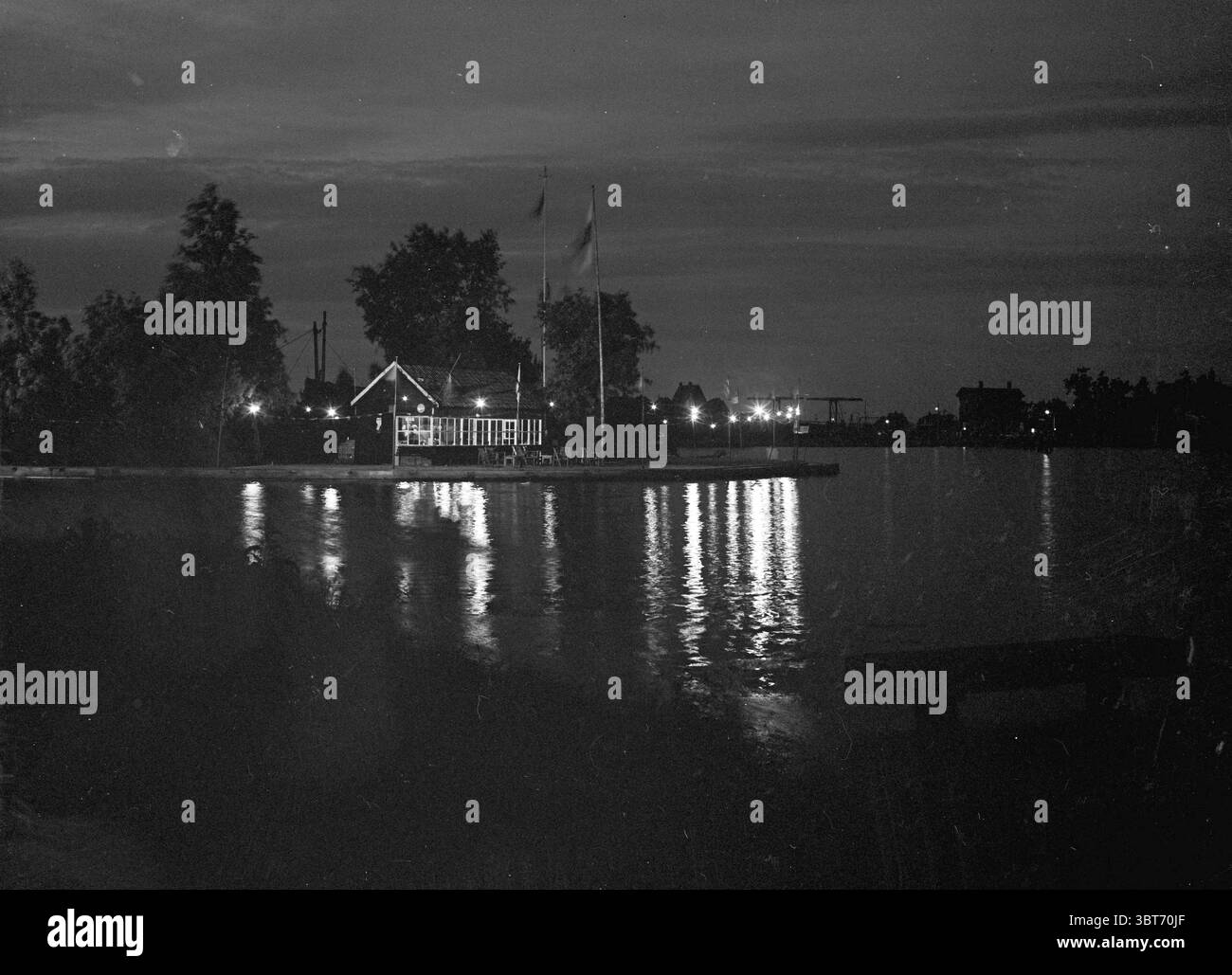 Yacht Shipyard de Rietpol à Spaarndam 15 ans anniversaire anniversaire anniversaires éclairage Spaarndam Rietpol 1, Whizgle News, Dutch Desk, pays-Bas, 1950 - 2000 on 01-06-1952. L'image montre ces rubriques. La scène capture un paysage nocturne serein, dominé principalement par des nuances de noir et blanc, évoquant une atmosphère nostalgique. Au premier plan, la surface calme de l'eau reflète de faibles lueurs de lumière, créant un chemin chatoyant qui attire le regard vers la rive opposée. D'un côté, une charmante structure semblable à une cabine est éclairée, ses fenêtres brillent d'une lueur chaleureuse Banque D'Images