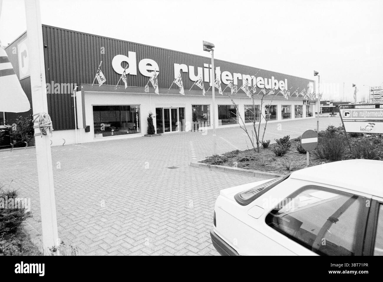 Magasin de meubles de Ruitermeubel fermé Cruquius, Whizgle News, Dutch Desk, The Netherlands, 1950 - 2000 le 24-12-1989. Ce sont les éléments de l'image. La scène présente un grand bâtiment moderne de vente au détail, caractérisé par sa façade expansive avec une combinaison de couleurs sombres et claires. La structure est dominée par de grandes fenêtres qui permettent à la lumière naturelle de filtrer, bien que l'atmosphère générale soit tamisée, comme l'indique le ciel couvert en arrière-plan. Des drapeaux de divers motifs flottent sur le toit du bâtiment et le long des côtés, ajoutant des éclaboussures de couleur contre les tons sourds de t Banque D'Images