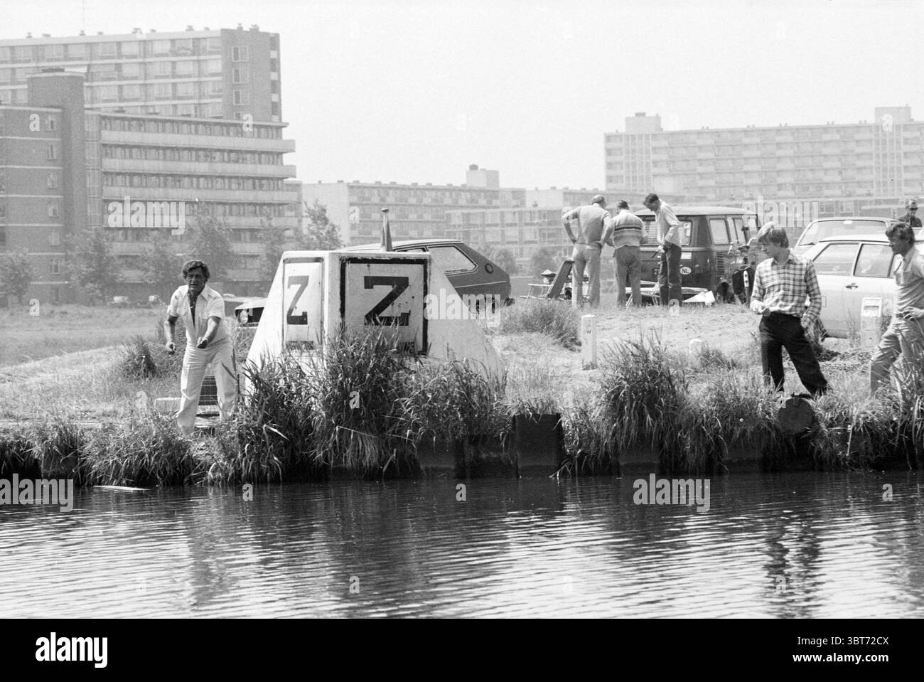 Pêche Wao'ers pêcheries et autres pêcheurs pêche, Whizgle News, Dutch Desk, pays-Bas, 1950 - 2000 le 05-07-1979. L'image montre ces rubriques. La scène capture un front de mer urbain animé par temps clair, avec la lumière du soleil illuminant la zone. Au premier plan, un petit groupe d'individus est engagé dans diverses activités près du bord de l'eau. Une personne, vêtue de vêtements légers, se tient à côté d'une grande structure en béton marquée de deux lettres proéminentes. Ils semblent examiner quelque chose dans l'eau. À proximité, un autre individu, portant une chemise à carreaux, regarde également Banque D'Images