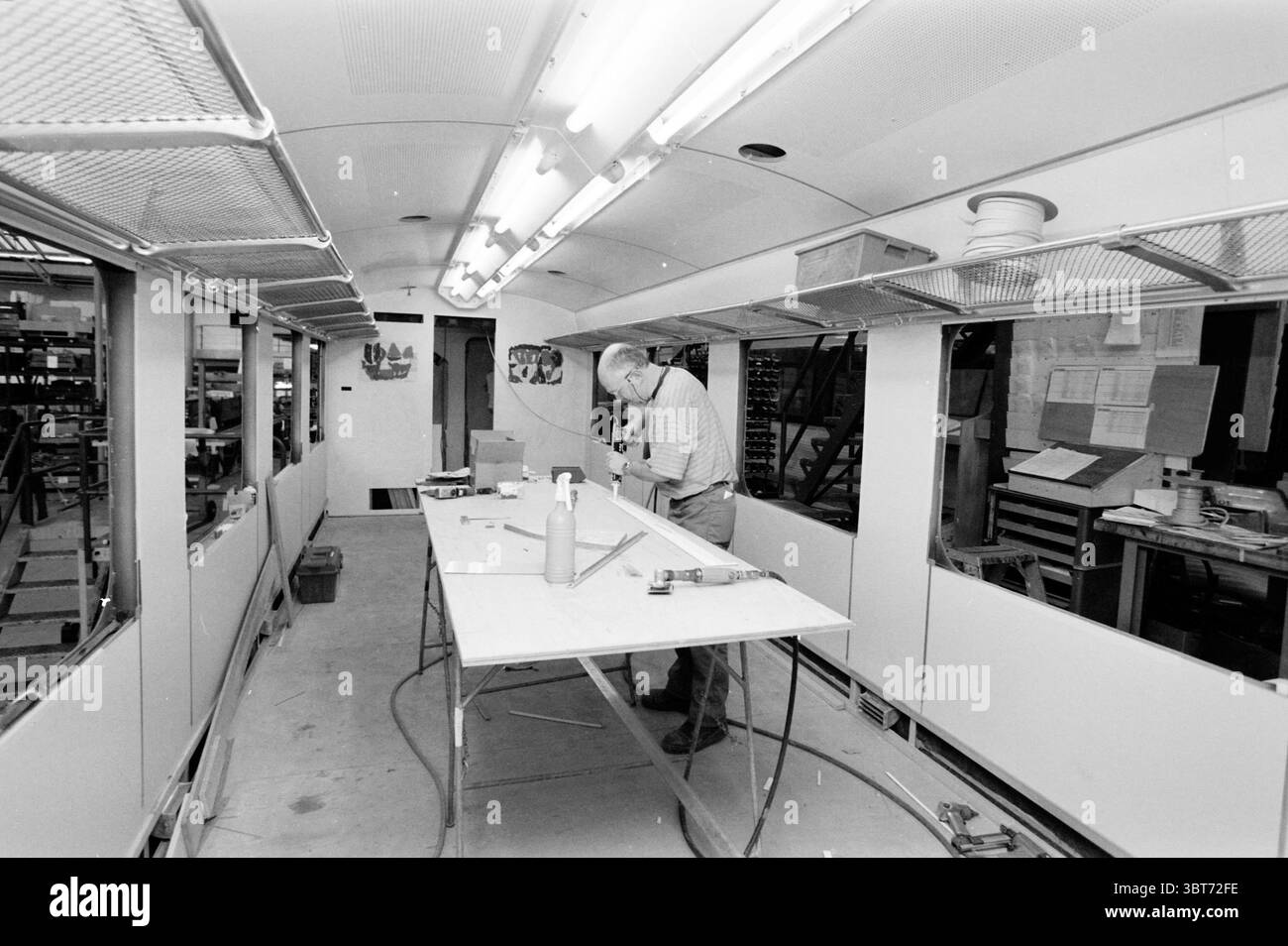 Build Burnt Out train, Whizgle News, Dutch Desk, pays-Bas, 1950 - 2000 le 08-02-2001. Ce sont les éléments de l'image. La scène représente un atelier intérieur, caractérisé par un espace long et étroit éclairé par des lumières fluorescentes brillantes au-dessus de la tête. Les murs sont majoritairement blancs, donnant à l'environnement un aspect propre et organisé, tandis que le sol a une finition gris utilitaire. Au centre, une grande table robuste occupe l'espace, encombrée de divers outils et matériaux. Il y a des éléments tels qu'un rouleau de ruban adhésif, un ensemble d'instruments de mesure, et des longueurs de bois, suggérant un Banque D'Images