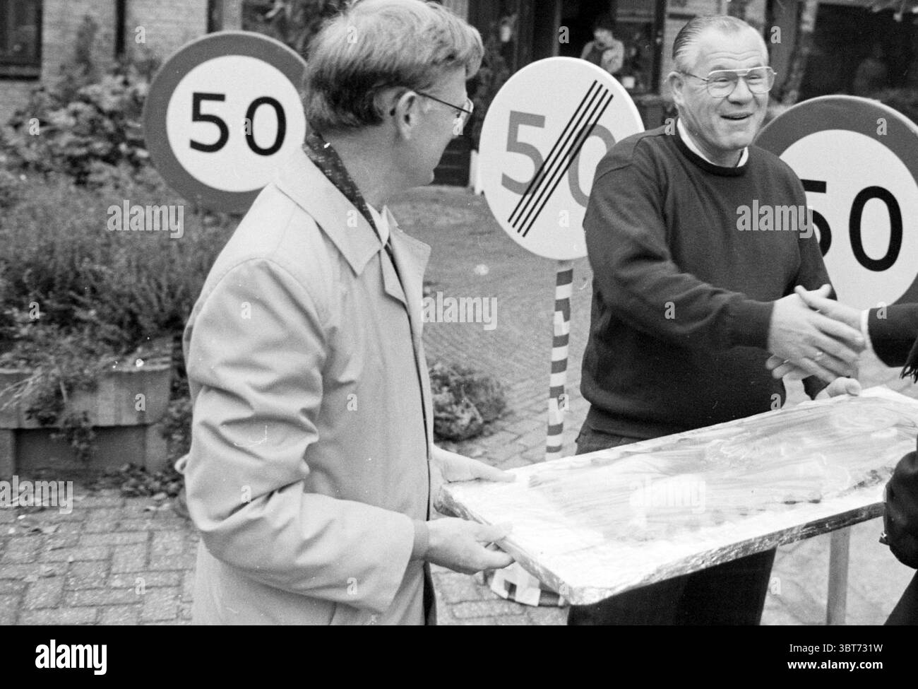 Alderman Auer Hoofddorp 50 ans anniversaires Aldermen Hoofddorp pays-Bas, Whizgle News, Dutch Desk, pays-Bas, 1950 - 2000 on 11-10-1988. Voici les rubriques de l'image. Dans un cadre extérieur animé, deux individus sont engagés dans un échange convivial. Un homme est habillé d'un manteau de couleur claire, tenant un grand plateau recouvert de ce qui semble être une surface brillante, affichant probablement une sorte de nourriture ou de dessert. Il a des lunettes et un soupçon de sourire, indiquant un comportement chaleureux. L'autre homme, portant un pull sombre, est en train de serrer la main avec le premier homme, mettant en vedette un Banque D'Images