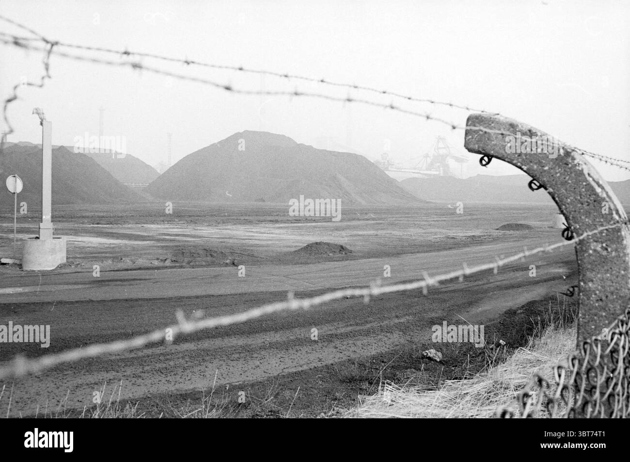 Montagnes de charbon de Hoogovens Hoogovens Velsen-Noord, Whizgle News, Dutch Desk, pays-Bas, 1950 - 2000 le 25-02-1982. L'image montre ces rubriques. La scène est dominée par un paysage industriel sombre, caractérisé par de grands monticules de sol sombre ou de gravier qui s'élèvent bien en évidence sur la toile de fond. Les tons sont atténués, principalement dans les nuances de gris, créant une atmosphère sombre et quelque peu désolée. Une clôture en fil de fer barbelé traverse le premier plan, son métal tranchant et torsadé contrastant fortement avec la terre du sol environnant. Cette barrière ajoute un sentiment de confinement ou de séparation Banque D'Images