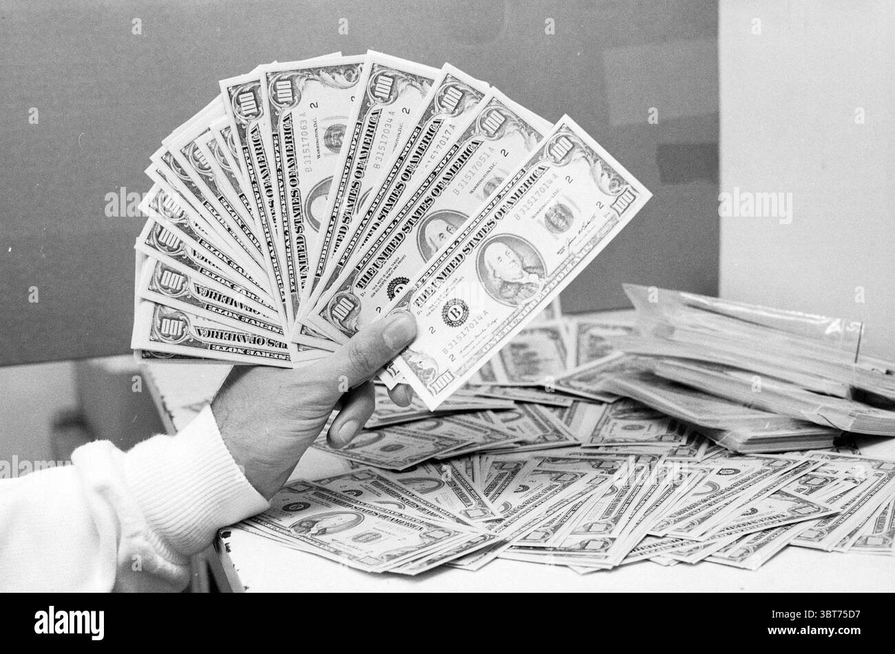 Faux billets de 100 dollars poste de police argent, Whizgle News, Dutch Desk, pays-Bas, 1950 - 2000 on 03-06-1987. Ces rubriques apparaissent dans l'image. Dans la scène, une main s'étend vers l'avant, affichant un éventail de devises. Les billets, principalement dans les tons de vert et de gris, sont soigneusement disposés, mettant en valeur diverses dénominations en évidence, avec les portraits et signatures emblématiques formant une toile de fond. La texture de la monnaie est nette, suggérant une manipulation récente. Derrière la main, il y a une surface encombrée de piles supplémentaires de ces billets, créant un sentiment d'abondance. L'arrière-plan apparaît bl Banque D'Images