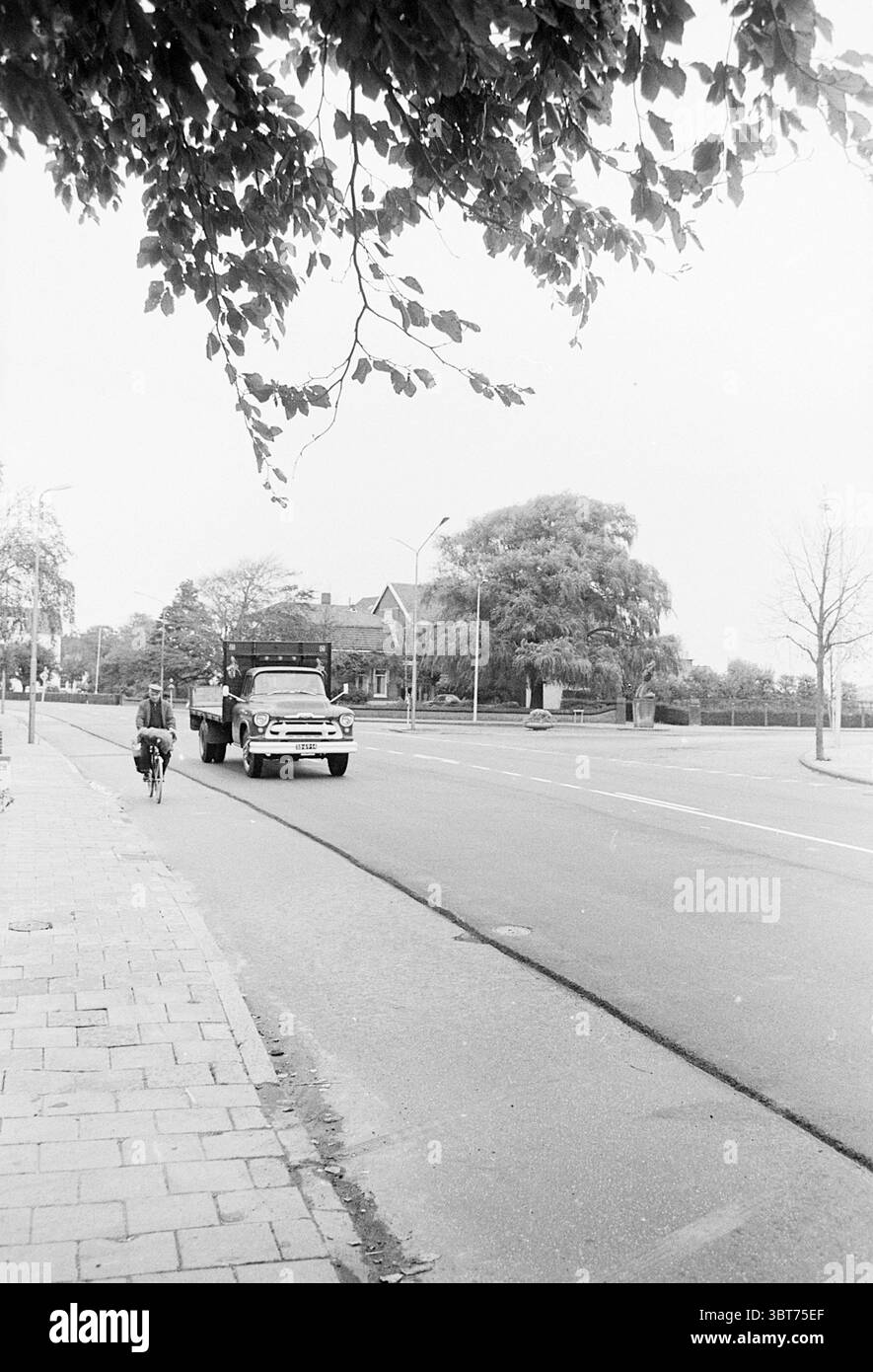 Jonction en T avec piste cyclable., Whizgle News, Dutch Desk, pays-Bas, 1950 - 2000. Voici les rubriques de l'image. La scène capture une rue calme, principalement ombragée par des arbres avec de larges branches verdoyantes. La route s'étend vers l'avant, révélant une surface lisse qui scintille légèrement, peut-être à cause des pluies récentes. À gauche, un cycliste est vu pédaler le long du trottoir, leur posture détendue mais déterminée. Le cycliste, vêtu d'une tenue décontractée, contraste avec les tons de gris plus discrets de l'environnement. Sur la route, un camion passe devant, ses accents chromés captent la lumière. Le tr Banque D'Images