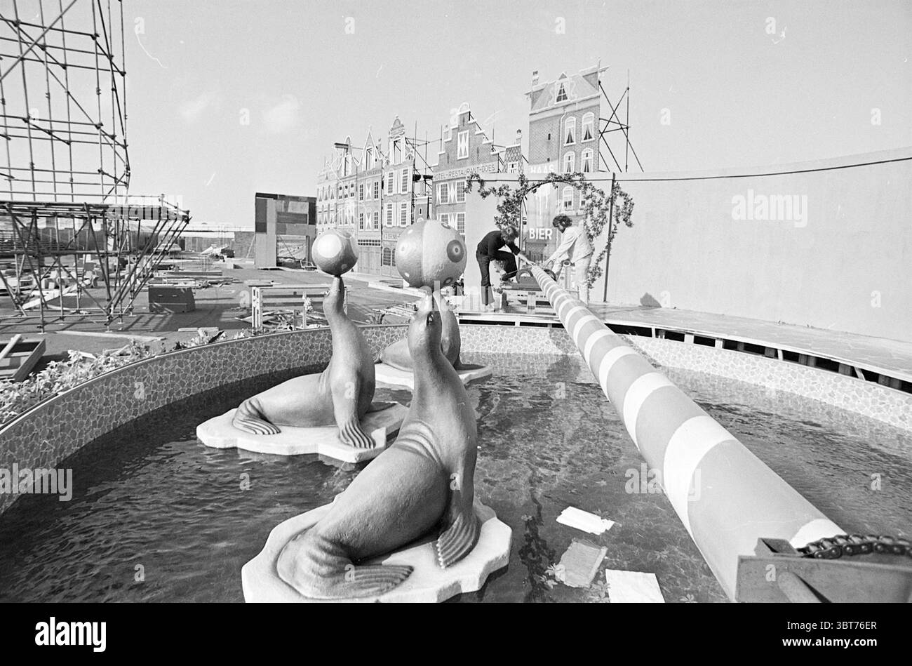 Construction d'un jeu sans frontières Zandvoort. Chantiers de construction, Whizgle News, Dutch Desk, pays-Bas, 1950 - 2000 sur 07-06-1974. L'image inclut ces rubriques. La scène capture une zone de construction qui mélange des éléments ludiques avec le développement continu. À l'avant-garde, une piscine peu profonde présente des sculptures fantaisistes de phoques, chacun équilibrant une sphère sur son nez. Les joints sont positionnés sur des plates-formes surélevées qui imitent les roches, et l'eau ondule doucement autour d'eux, créant une sensation de plaisir. En arrière-plan, une série de bâtiments imitent l'architecture européenne traditionnelle, cha Banque D'Images