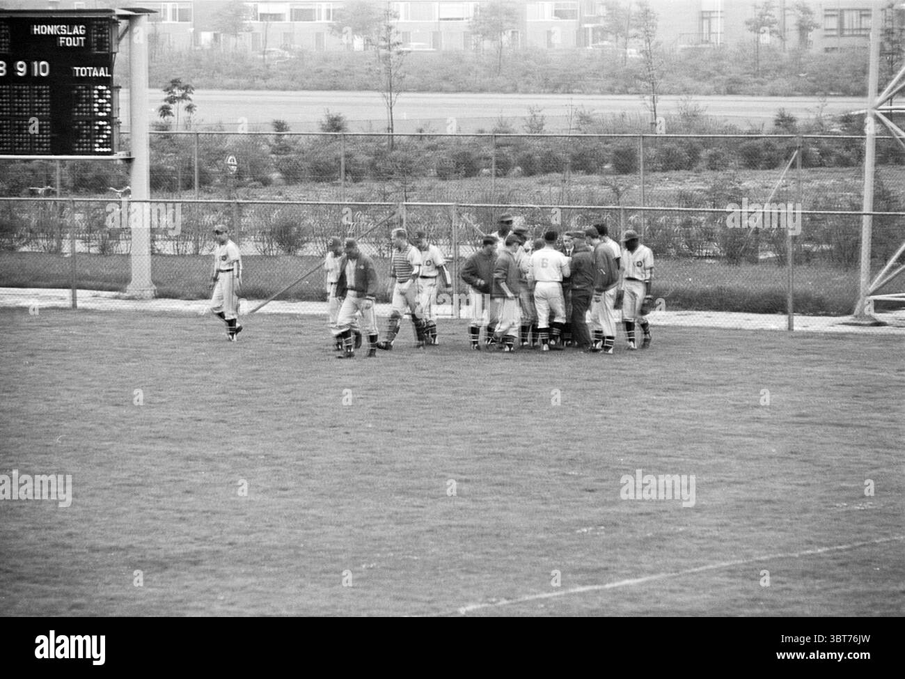 Coups de feu - Sparta Baseball, Whizgle News, Dutch Desk, pays-Bas, 1950 - 2000 sur 18-05-1964. L'image inclut ces rubriques. La scène capture un moment sur un terrain de sport, probablement un match de baseball ou de softball. Au premier plan, un groupe de joueurs est entassé, leurs uniformes suggérant un esprit d'équipe cohésif. Ils présentent une gamme de postures, certaines se penchant étroitement tandis que d'autres se tiennent légèrement à l'écart, indiquant un moment de communication ou de discussion stratégique. Les joueurs portent des uniformes de couleur claire avec des accents sombres, probablement conçus pour fournir de la clarté contre le terrain vert luxuriant ben Banque D'Images