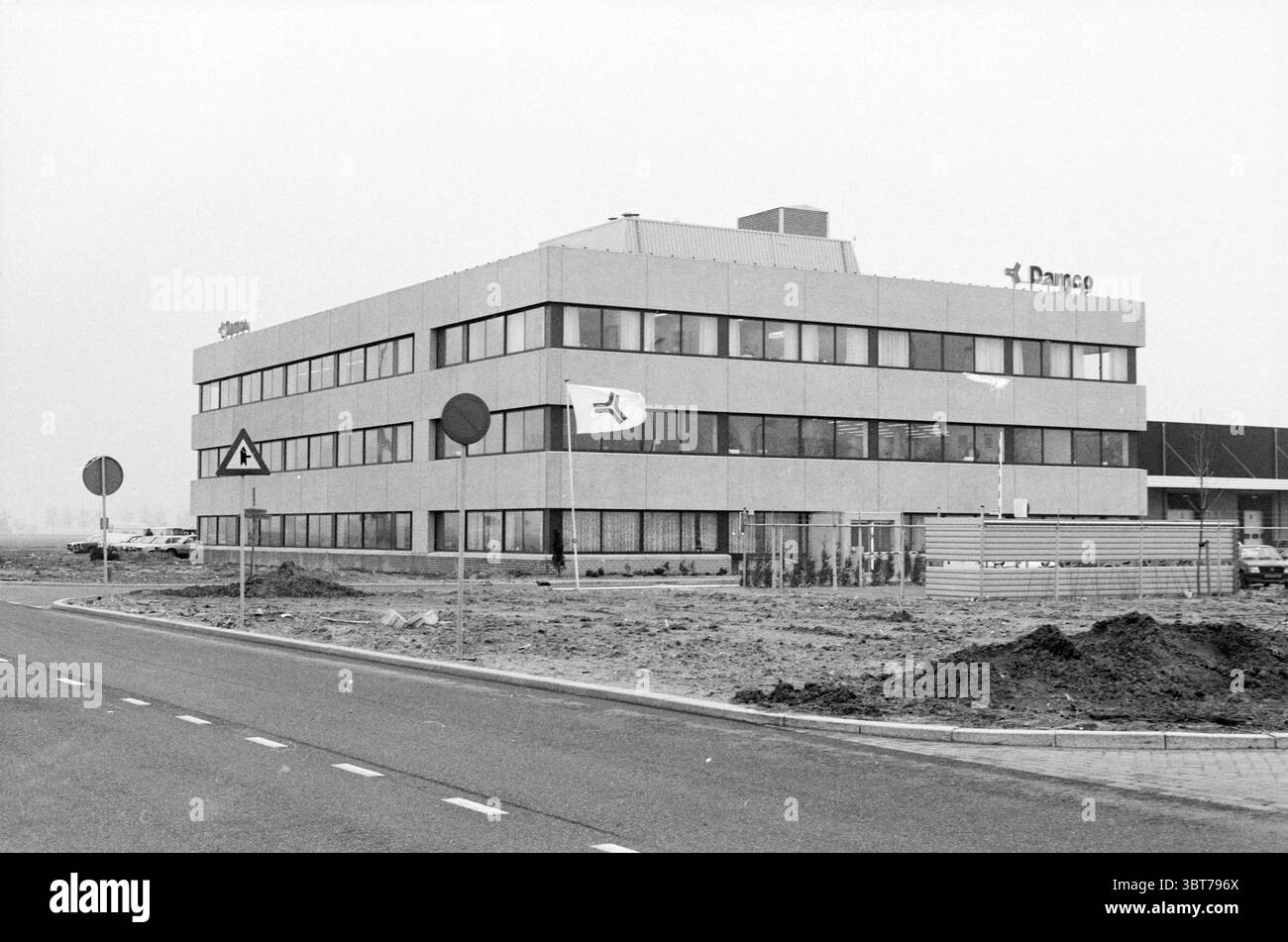 Immeuble de bureaux extérieur de Damco., Whizgle News, Dutch Desk, pays-Bas, 1950 - 2000. Voici les rubriques de l'image. La scène présente un bâtiment moderne à plusieurs niveaux caractérisé par son design géométrique et une combinaison d'éléments en béton et en verre. La structure a une esthétique minimaliste, avec un toit plat orné d'une petite saillie centrale. Situé au bord d'un paysage aride, l'extérieur du bâtiment est principalement gris, créant un contraste frappant avec le ciel plus clair au-dessus. Devant le bâtiment, une route bien entretenue courbe doucement, bordée d'une bordure o soignée Banque D'Images