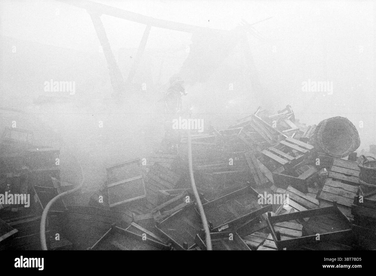 Hangar à ampoules à feu fa. Willemsen Hillegom Fire brigade Hillegom, Whizgle News, Dutch Desk, pays-Bas, 1950 - 2000 on 02-03-1980. Ces rubriques apparaissent dans l'image. La scène dépeint une atmosphère chaotique et dense, remplie d'une brume épaisse et grisâtre qui enveloppe l'environnement, faisant allusion à un incendie récent ou en cours. Au premier plan, un nombre considérable de caisses en bois sont empilées de manière désordonnée, certaines renversées, suggérant une scène de désarroi et d'urgence. Émergeant du brouillard, un personnage, probablement un pompier, est partiellement visible, vêtu d'un équipement de protection, tenant un long tuyau qui Banque D'Images