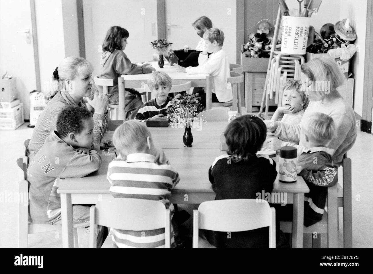 Babushka Childcare Haarlem pays-Bas, Whizgle News, Dutch Desk, pays-Bas, 1950 - 2000 on 10-06-1999. L'image inclut ces rubriques. La scène capture un cadre intérieur animé, probablement une salle de classe ou un espace commun conçu pour les enfants. L'espace est rempli d'une lumière douce et diffuse qui crée une atmosphère chaleureuse et accueillante. Au centre, une table rectangulaire en bois clair est entourée de plusieurs enfants, chacun engagé dans une conversation tranquille ou concentré sur leurs activités. Les enfants affichent une variété de couleurs et de styles de cheveux, contribuant à un sentiment de diversité. Banque D'Images