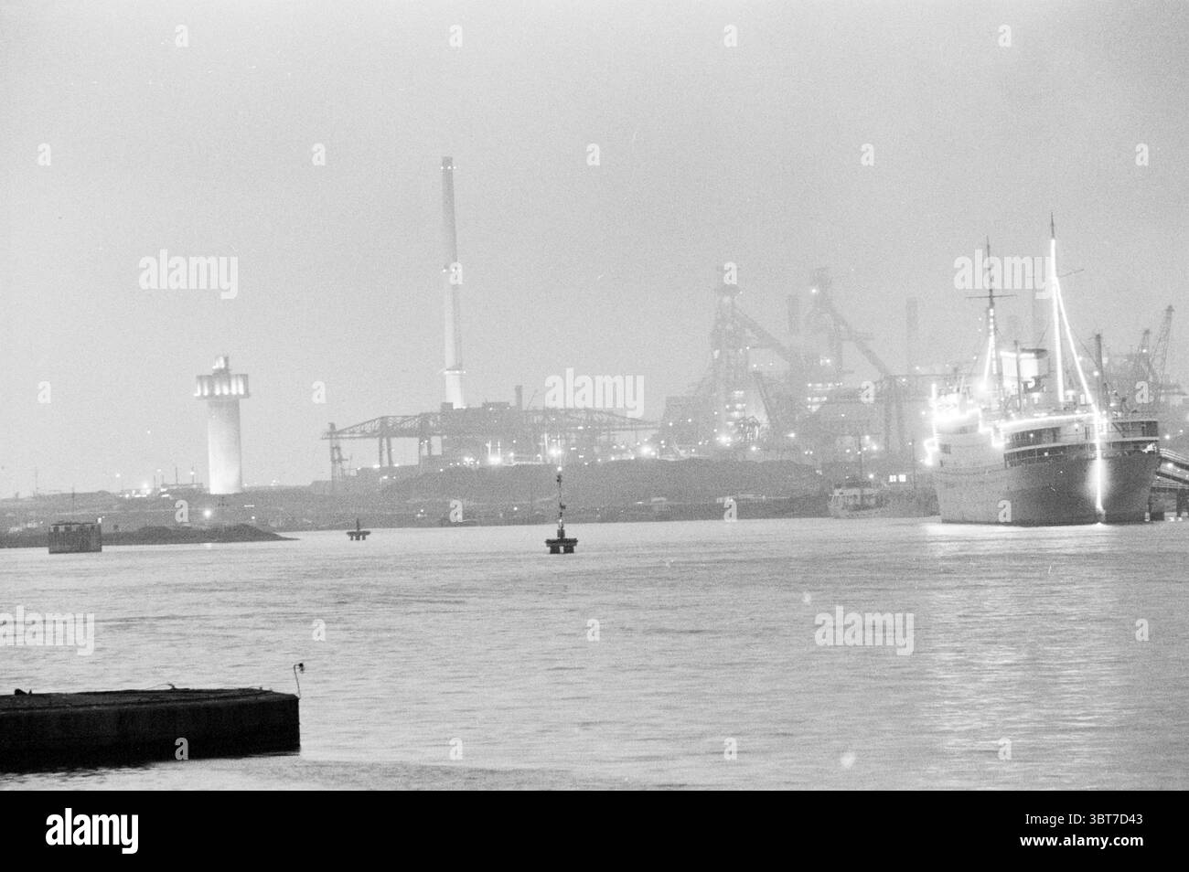Hauts fourneaux IJmuiden IJmuiden pays-Bas., Whizgle News, Dutch Desk, pays-Bas, 1950 - 2000. Ces rubriques sont illustrées dans l'image. La scène capture un port entouré d'une brume fantomatique voilée, créant une atmosphère de mystère tranquille. Au premier plan, l'eau sombre reflète un scintillement discret, suggérant une surface calme qui déforme les contours des objets au-dessus. A droite, un grand navire est amarré, sa silhouette frappée par le brouillard plus léger. Le navire a un design classique et robuste, avec des détails proéminents tels qu'une grande cheminée et des structures de pont. Sa coque semble sombre et Banque D'Images