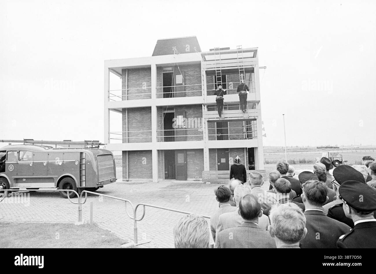 Ouverture de la tour de pompiers pompiers pompiers, Whizgle News, Dutch Desk, pays-Bas, 1950 - 2000 le 02-07-1965. L'image montre ces rubriques. La scène capture une journée nuageuse avec un ciel couvert et silencieux, contribuant à une atmosphère quelque peu tamisée. Au centre se dresse une structure de bâtiment moderne, caractérisée par ses lignes épurées et son design minimaliste. Il se compose de plusieurs niveaux, avec une façade en béton et de grandes fenêtres qui reflètent les tons gris de l'environnement. Deux figures en uniformes sont visibles, positionnées sur un échafaudage au niveau supérieur du bâtiment. Ils semblent s'y engager Banque D'Images