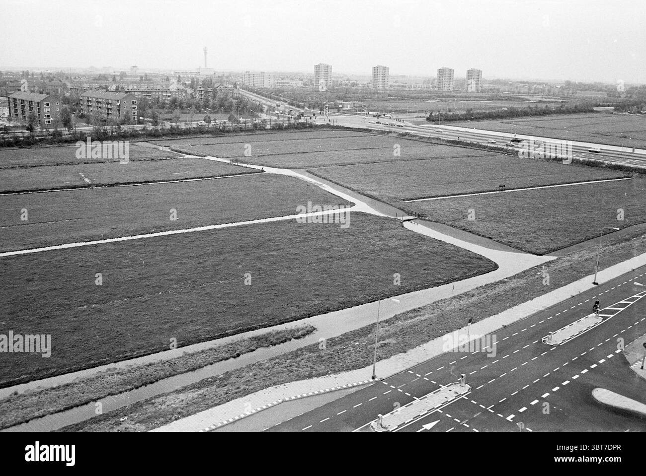 Panorama main Road., Whizgle News, Dutch Desk, pays-Bas, 1950 - 2000. L'image montre ces rubriques. La scène présente une vue large et expansive d'un paysage marqué par des champs plats et cultivés. Le sol est divisé en parcelles rectangulaires de verdure, probablement des terres agricoles, séparées par des voies navigables étroites et sinueuses qui brillent faiblement. Ces caractéristiques de l'eau créent un motif fluide et courbe sur le terrain, contrastant avec les lignes rigides de la culture. En arrière-plan, une série de bâtiments de faible hauteur se dessinent, leurs façades simples et géométriques. Leurs tons sourds se fondent dans le ciel, ce qui est o Banque D'Images