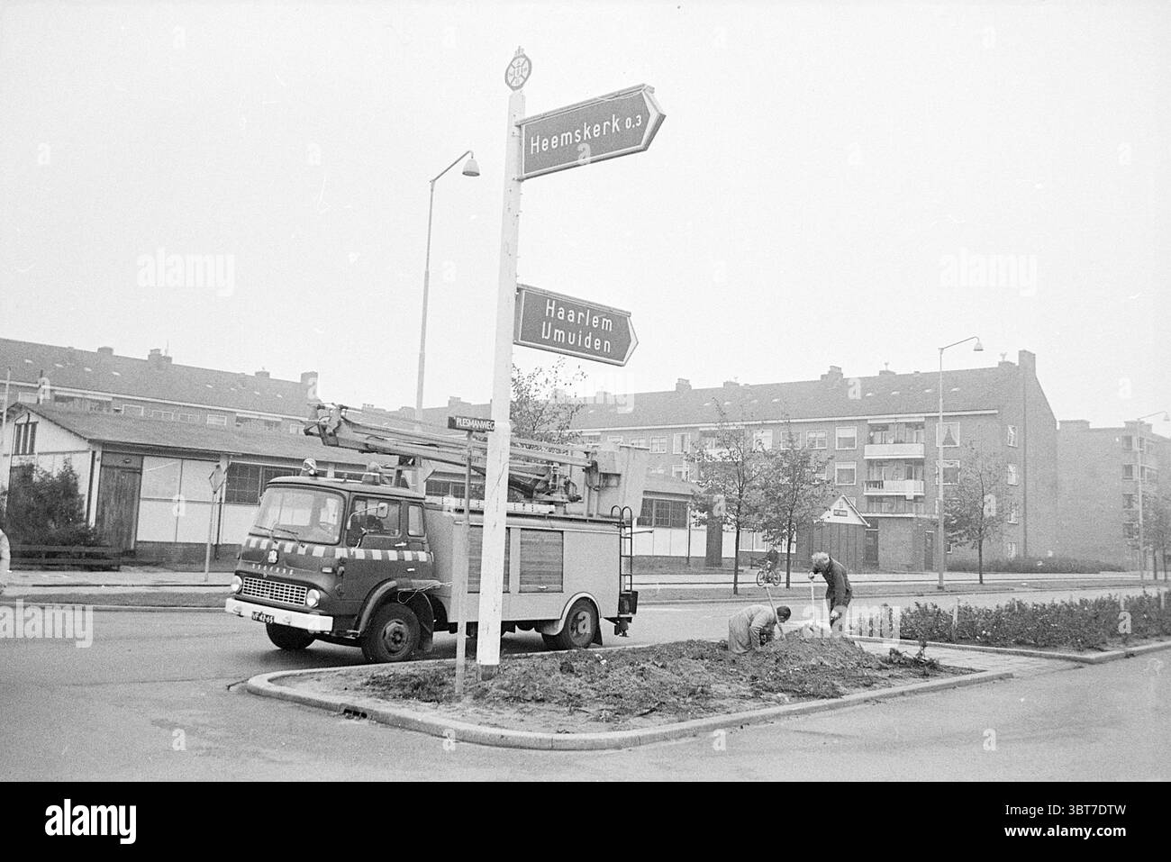 ANWB signe Beverwijk Signs, Whizgle News, Dutch Desk, pays-Bas, 1950 - 2000 le 08-10-1970. L'image montre ces rubriques. La scène capture un paysage urbain silencieux sous un ciel couvert, créant une atmosphère sombre mais engageante. Un camion de pompiers vintage, peint dans une nuance de gris terne avec des marques distinctives, est stationné bien en évidence à gauche. Son échelle étendue, il suggère la disponibilité à l'action, donnant un sens de l'objectif fonctionnel au véhicule. Au premier plan, deux individus sont agenouillés sur le sol, engagés dans des activités qui semblent impliquer le jardinage ou l'entretien. Le Banque D'Images
