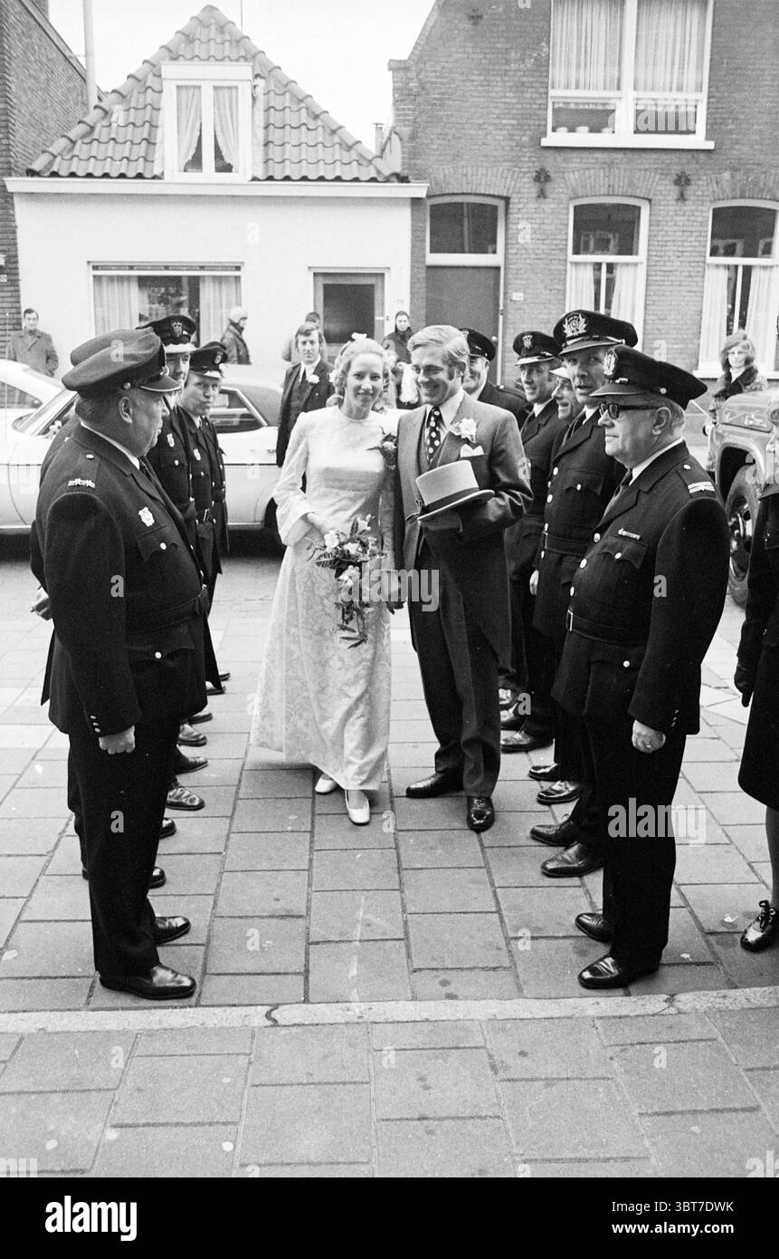 Mariage des pompiers., Whizgle News, Dutch Desk, pays-Bas, 1950 - 2000. Ces rubriques sont illustrées dans l'image. Dans une scène animée en plein air, un couple joyeux se tient au premier plan, rayonnant de joie et de célébration. La mariée, vêtue d'une élégante robe blanche avec une délicate superposition de dentelle, tient un bouquet de fleurs fraîches, ce qui ajoute une touche vibrante de couleur à son ensemble. Ses cheveux sont magnifiquement coiffés, mettant en valeur des vagues douces qui encadrent son visage. À côté d'elle, le marié porte un costume pointu et sombre complété par une chemise de couleur claire et une cravate, ce qui rehausse l'atmosphère formelle mais festive. T Banque D'Images