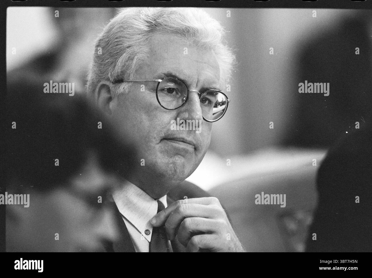 Conseillers budgétaires H'lem Haarlem Grote Markt 2 pays-Bas, Whizgle News, Dutch Desk, pays-Bas, 1950 - 2000 on 03-11-1996. L'image montre ces rubriques. La scène capture un homme assis dans un rassemblement, son expression réfléchie et légèrement pensive. Il a les cheveux grisonnés coiffés soigneusement, encadrant son visage, qui porte des notes d'âge avec des lignes fines autour de ses yeux. Les lunettes rondes encadrées en métal accentuent son regard concentré. Sa tenue est formelle, avec un costume de couleur claire et une cravate qui suggère professionnalisme et sérieux. Une main est légèrement relevée, les doigts reposant sur son c Banque D'Images