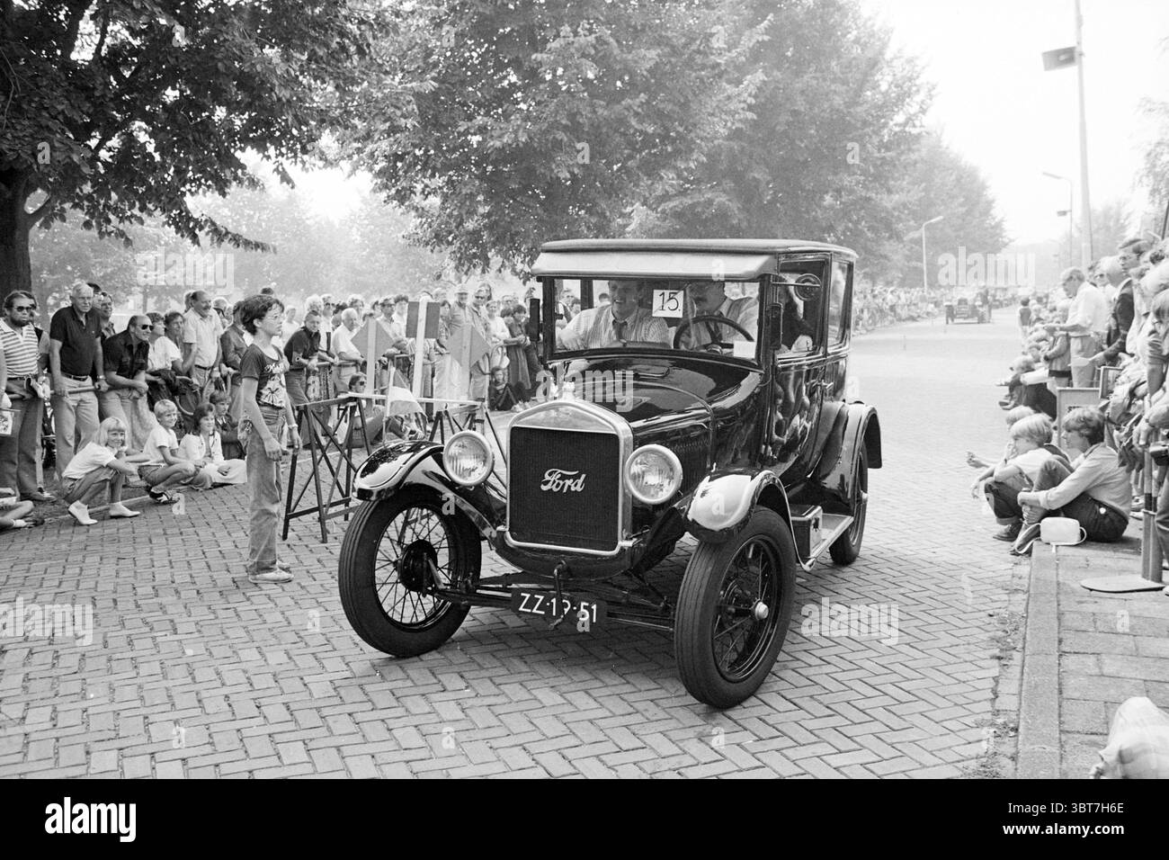 Procession de voitures classiques, Whizgle News, Dutch Desk, pays-Bas, 1950 - 2000 le 06-08-1982. L'image inclut ces rubriques. La scène capture un événement de rue animé qui rappelle une époque révolue, marquée par une automobile noire classique qui attire l'attention sur un chemin pavé. Ce véhicule vintage, caractérisé par ses courbes lisses et ses détails distinctifs, est mis en évidence au centre de la composition, mettant en valeur son charme nostalgique. Autour de la voiture, une foule de spectateurs crée un sentiment d'excitation et d'engagement. Les gens varient en apparence et en tenue vestimentaire, Banque D'Images