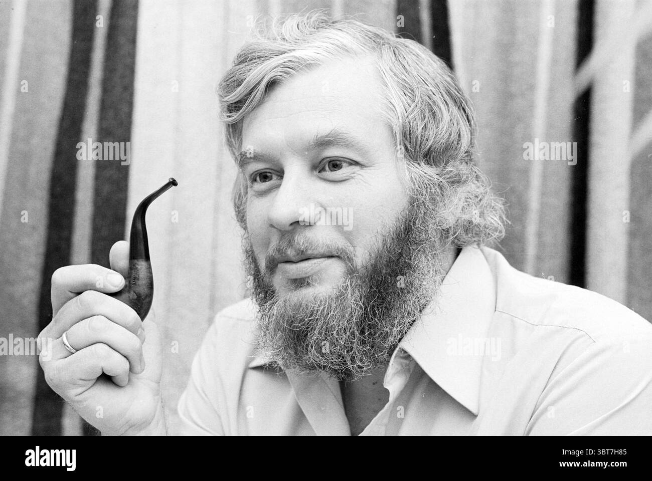 Portrait Mr. Steyn IJmuiden Portraits de police IJmuiden pays-Bas, Whizgle News, Dutch Desk, pays-Bas, 1950 - 2000 on 07-08-1980. L'image inclut ces rubriques. Un homme à la barbe épaisse et pleine et à la coiffure rappelant les années 1970 est assis dans une position détendue, dégageant un sentiment de calme et de contemplation. Il tient délicatement entre ses doigts une pipe à tabac courbée et sombre, la surface lisse de la pipe contrastant avec la texture de sa barbe. Sa chemise est légèrement colorée, peut-être un pastel doux, complétant la palette chaude et discrète de l'arrière-plan, qui se compose de rayures verticales en va Banque D'Images