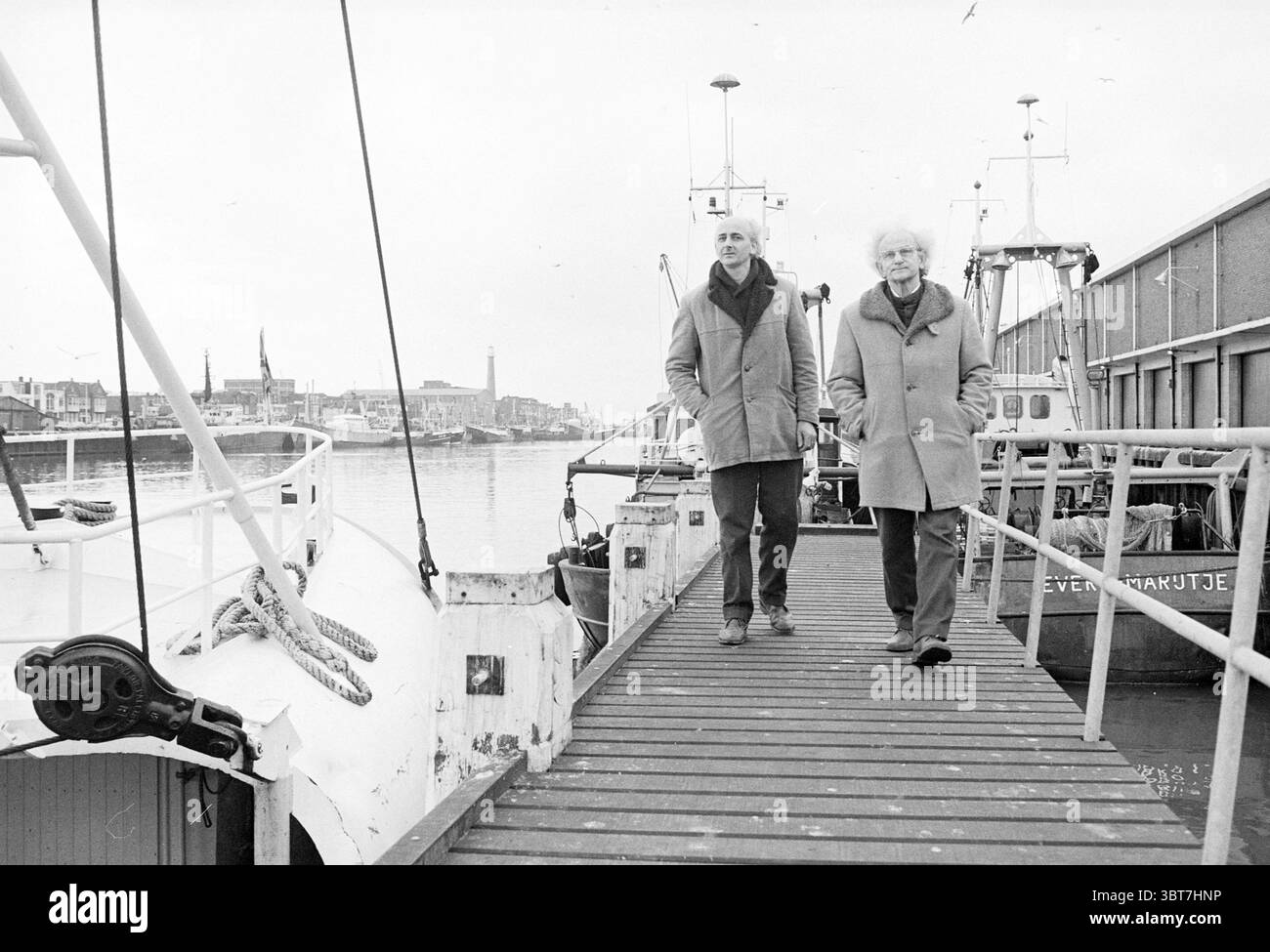Père et fils Van Woensel ont inventé des inventions de machine à éplucher les crevettes, Whizgle News, Dutch Desk, pays-Bas, 1950 - 2000 le 17-12-1978. Ces rubriques sont illustrées dans l'image. La scène capture un cadre portuaire monochrome, évoquant un sentiment de nostalgie et de tranquillité. Deux individus marchent le long d'un quai en bois, leurs personnages bien en vue sur la toile de fond des bateaux amarrés. La première personne présente un comportement distingué, portant un manteau de couleur claire, tandis que la seconde, avec des cheveux blancs tortueux, arbore un manteau plus foncé. Leur posture détendue suggère confort et familiarité avec chaque o Banque D'Images