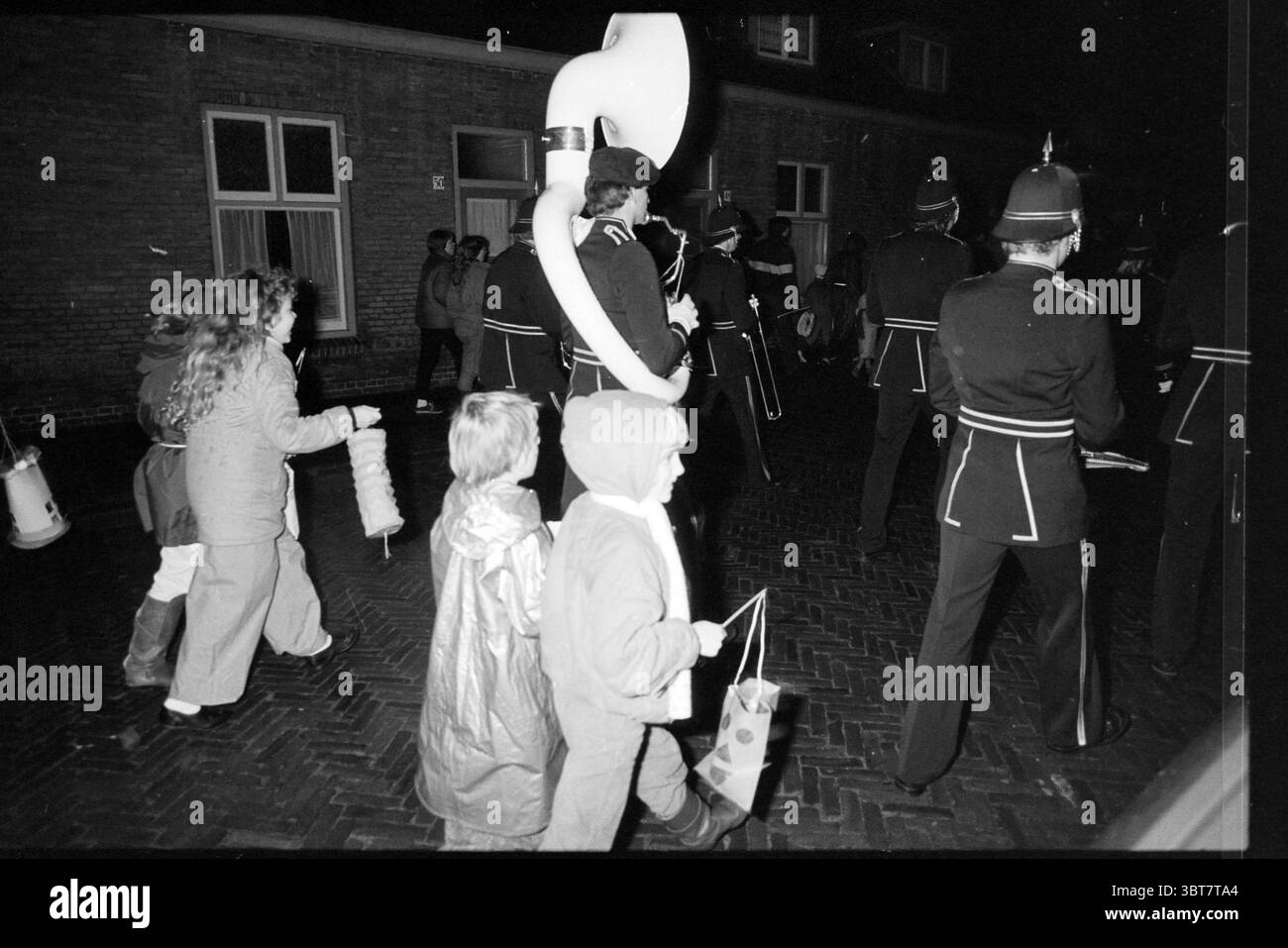 Sint Maarten parade Godfried Bouillonstraat composé Maarten Haarlem Godfried van Bouillonstraat pays-Bas, Whizgle News, Dutch Desk, pays-Bas, 1950 - 2000 on 11-11-1986. Ces rubriques apparaissent dans l'image. La scène se déroule la nuit avec un rassemblement de gens marchant le long d'une rue sombre et pavée. L'air est rempli d'un sentiment de fête et de communauté. Au premier plan, il y a plusieurs enfants. Un enfant est habillé d'une capuche floue, paraissant chaud et confortable, tandis qu'un autre arbore un imperméable brillant avec la capuche relevée, suggérant de l'excitation et peut-être un peu de méfait. Elles Banque D'Images