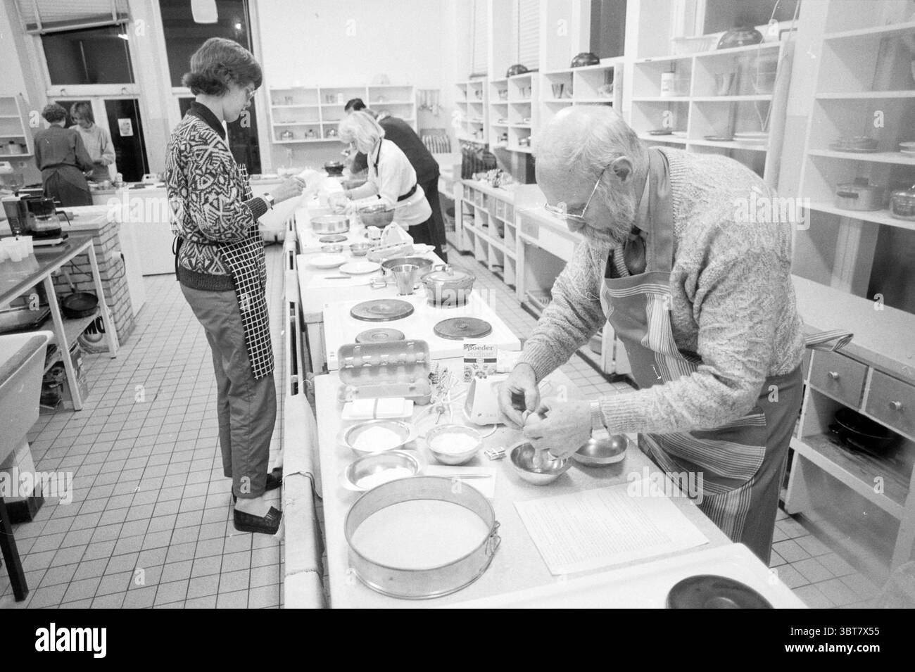 Cours de cuisine pour adultes Franciscusschool IJmuiden cuisine IJmuiden pays-Bas, Whizgle News, Dutch Desk, pays-Bas, 1950 - 2000 on 28-11-1988. Voici les rubriques de l'image. La scène capture un environnement de cuisine animé, rempli d'un sentiment de collaboration et de créativité. Au premier plan, un homme avec des lunettes est intentionnellement concentré sur sa tâche, portant un tablier sur un pull léger. Il mesure délicatement les ingrédients dans de petits bols, entourés de divers ustensiles de cuisine et de récipients qui font allusion à des activités de cuisson ou de cuisson. Son comportement suggère concentration et CA Banque D'Images