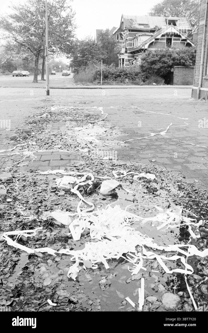 Paper on the Street Kanaalstraat IJmuiden Garbage IJmuiden Kanaalstraat pays-Bas, Whizgle News, Dutch Desk, pays-Bas, 1950 - 2000 sur 15-10-1987. Ces rubriques sont illustrées dans l'image. La scène présente un paysage urbain qui combine des éléments de la nature et de l’activité humaine. Au premier plan, un trottoir humide et inégal est parsemé de feuilles tombées, ajoutant une touche de chaleur à l'environnement autrement couvert. Parmi les feuilles se trouvent des touffes de matière blanche, peut-être du papier ou du plastique mis au rebut, contrastant fortement avec les tons terreux du sol humide. Une flaque d'eau scintille dans la lumière, Re Banque D'Images
