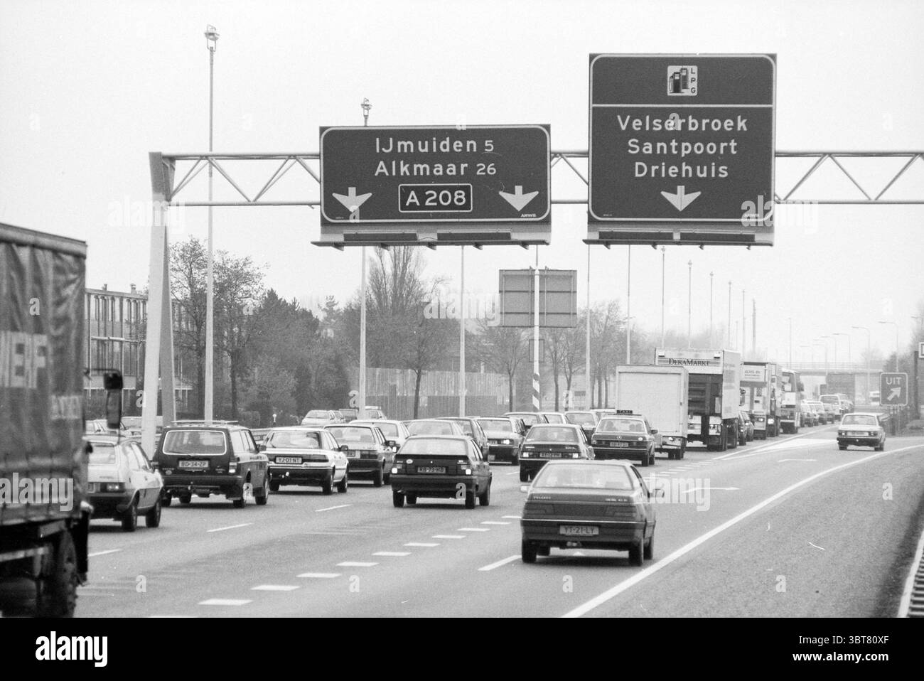 Embouteillage A208 près de Velserbroek Velserbroek, Whizgle News, Dutch Desk, pays-Bas, 1950 - 2000 le 02-04-1991. L'image inclut ces rubriques. La scène représente une route très fréquentée, caractérisée par une ligne continue de véhicules se déplaçant dans les deux sens. De nombreuses voitures peuvent être vues, principalement dans les tons gris, argent et noir, reflétant une atmosphère monochrome. Les véhicules sont très bien emballés, ce qui indique un état de congestion, certains étant à l'arrêt et d'autres se déplaçant lentement vers l'avant. Au-dessus de la chaussée, deux grands panneaux directifs sont suspendus, leurs fonds dans un vert légèrement délavé, affichant du blanc Banque D'Images