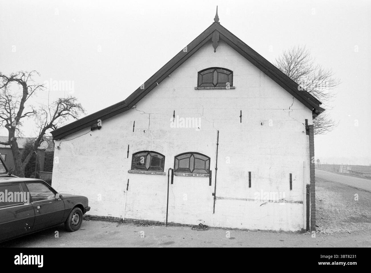 Old Forge de Kwakel de Kwakel, Whizgle News, Dutch Desk, pays-Bas, 1950 - 2000 on 12-12-1991. L'image contient ces rubriques. La scène représente un bâtiment modeste et légèrement usé avec un design simple qui respire un charme rustique. La structure est principalement blanchie à la chaux, bien que la peinture montre des signes d'altération et d'âge, caractérisés par des fissures et des taches de décoloration. Le toit est incliné avec une forme sombre et triangulaire, avec une petite flèche décorative au sommet qui ajoute une touche de caractère. Flanquant le bâtiment, il y a quelques arbres sans feuilles, leurs branches frappent fortement contre le Banque D'Images