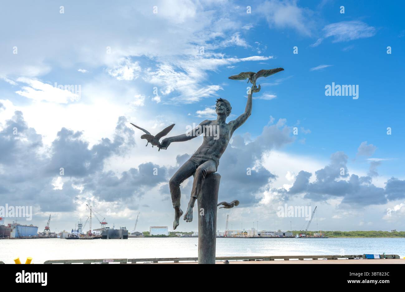Galveston, USA - 11 juillet 2025 : statue en bronze d'un garçon avec mouettes sur le front de mer avec ciel nuageux et vue sur le port Banque D'Images