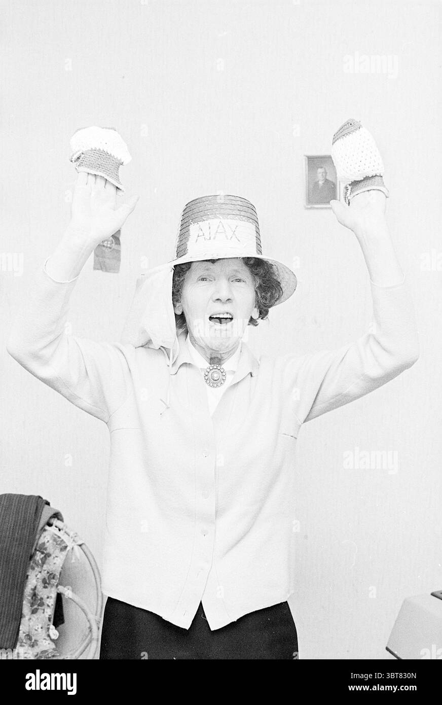 MRS van Vooren Ajax supporters Persons, Whizgle News, Dutch Desk, pays-Bas, 1950 - 2000 on 24-04-1969. L'image inclut ces rubriques. La scène met en scène une femme plus âgée expressive au centre, avec un comportement joyeux et animé. Elle porte un grand chapeau de paille orné d'une simple bande, qui attire le regard et ajoute à sa présence vibrante. Ses cheveux sont coiffés d'une manière qui suggère un esprit insouciant, encadrant son visage. Elle tient deux articles tricotés colorés dans chaque main, les élevant avec enthousiasme, peut-être dans le cadre d'une célébration ou d'une performance. Les articles semblent être ludiques et à la main Banque D'Images